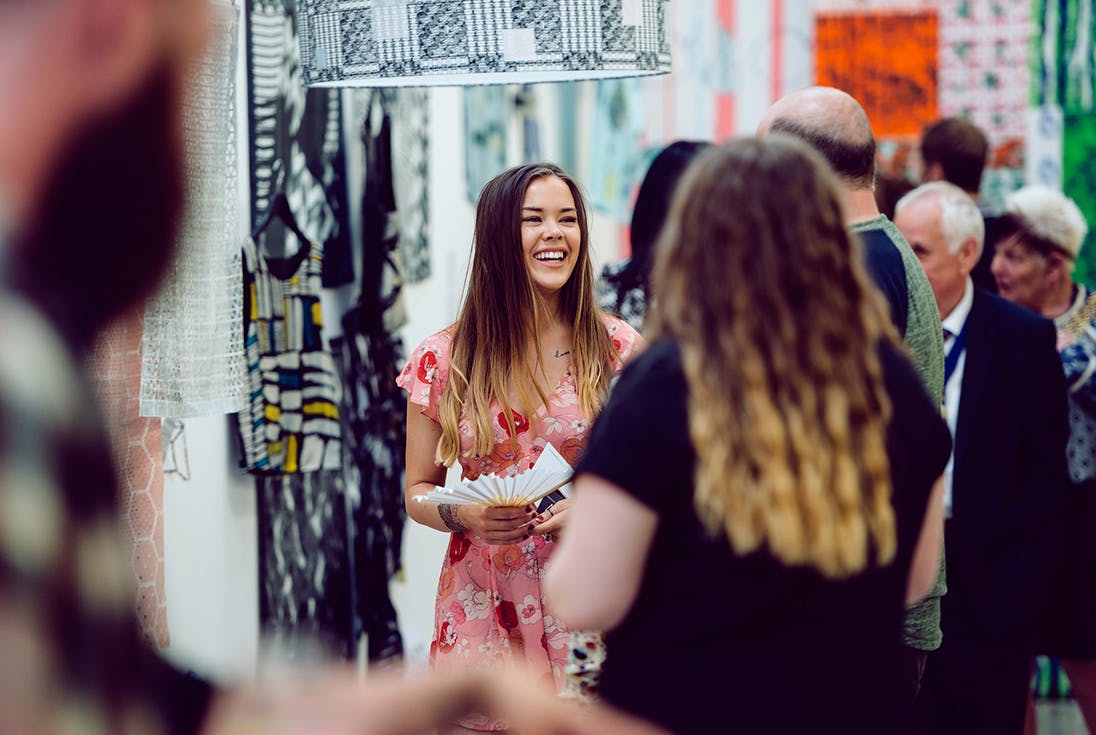 A happy smiling female student is surrounded by people at an exhibition with colourful textiles in the background