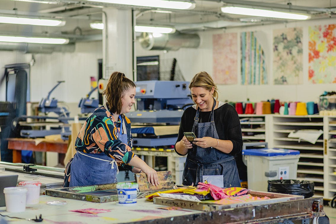 A smiling student screenprinting in the Textiles studios with technical demonstrator Kathryn Hays