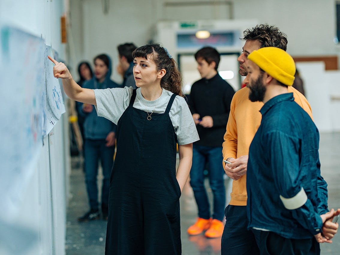 A woman wearing black dungarees points to drawings and notes on a wall whilst chatting to two men standing close by