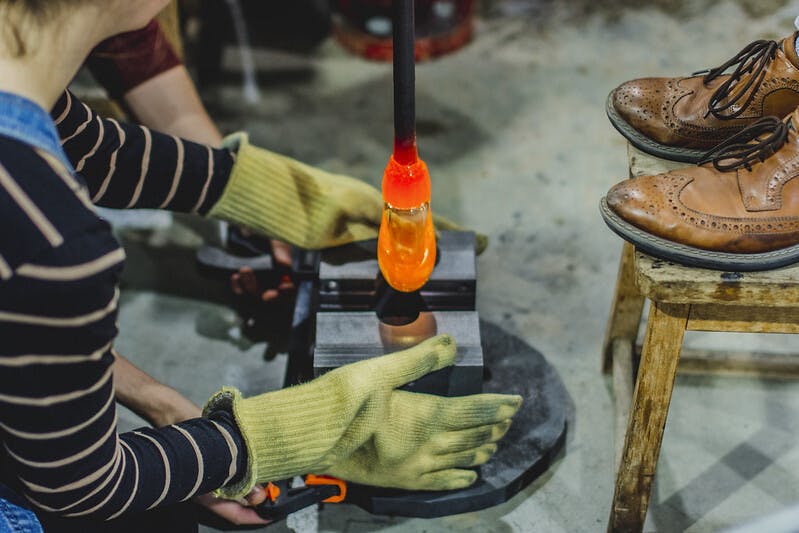 Glowing from the furnace, a hot orange blob of molten glass is carefully placed for shaping, the students wear protective gloves.