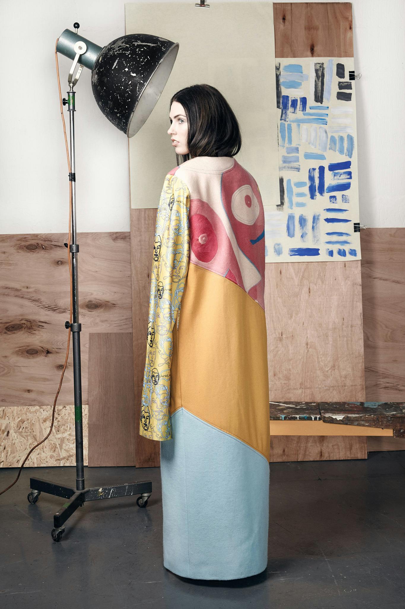 Poppy Yeoman's design from her 2016 graduation show. A female model with a sleek black bob, wears a long gown with yellow and blue panels. She is facing away from the camera, the top panel of the gown features a face-like design with patterned sleeves.