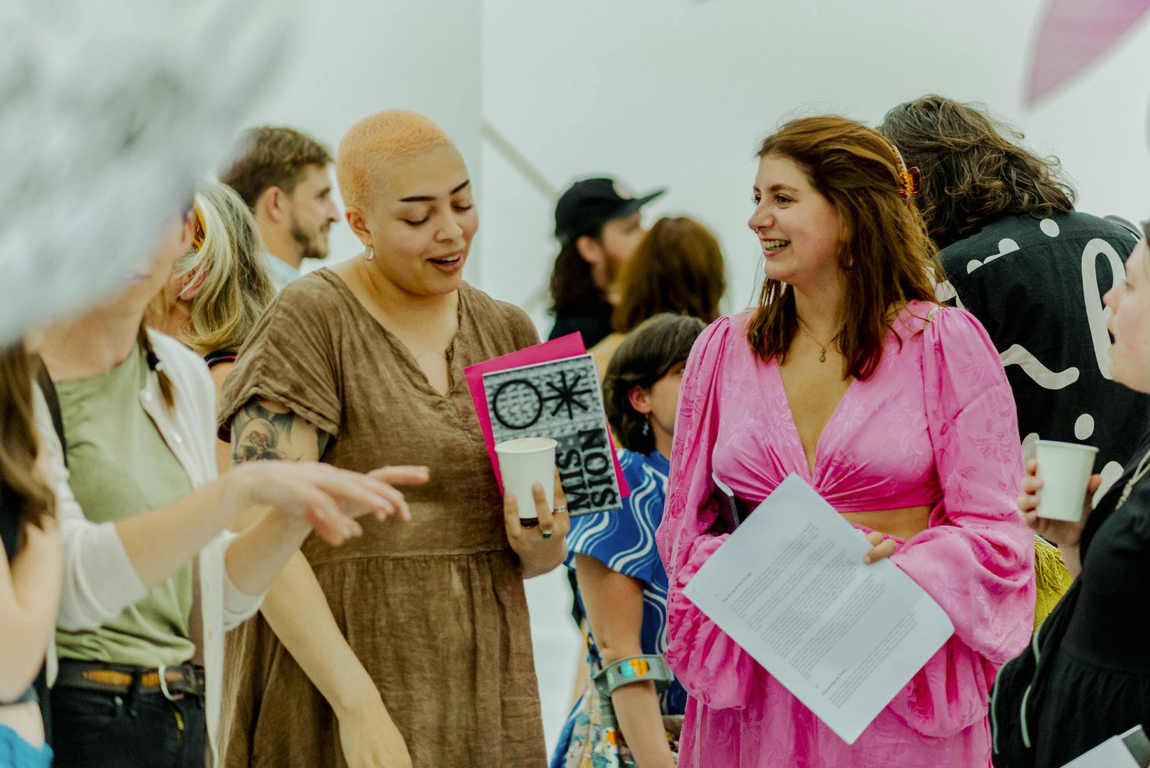 A person with short peach coloured hair stands next to a person in a pink dress amongst a crowd of people enjoying the Arts University Plymouth BA Hons Fine Art Show in June 2022