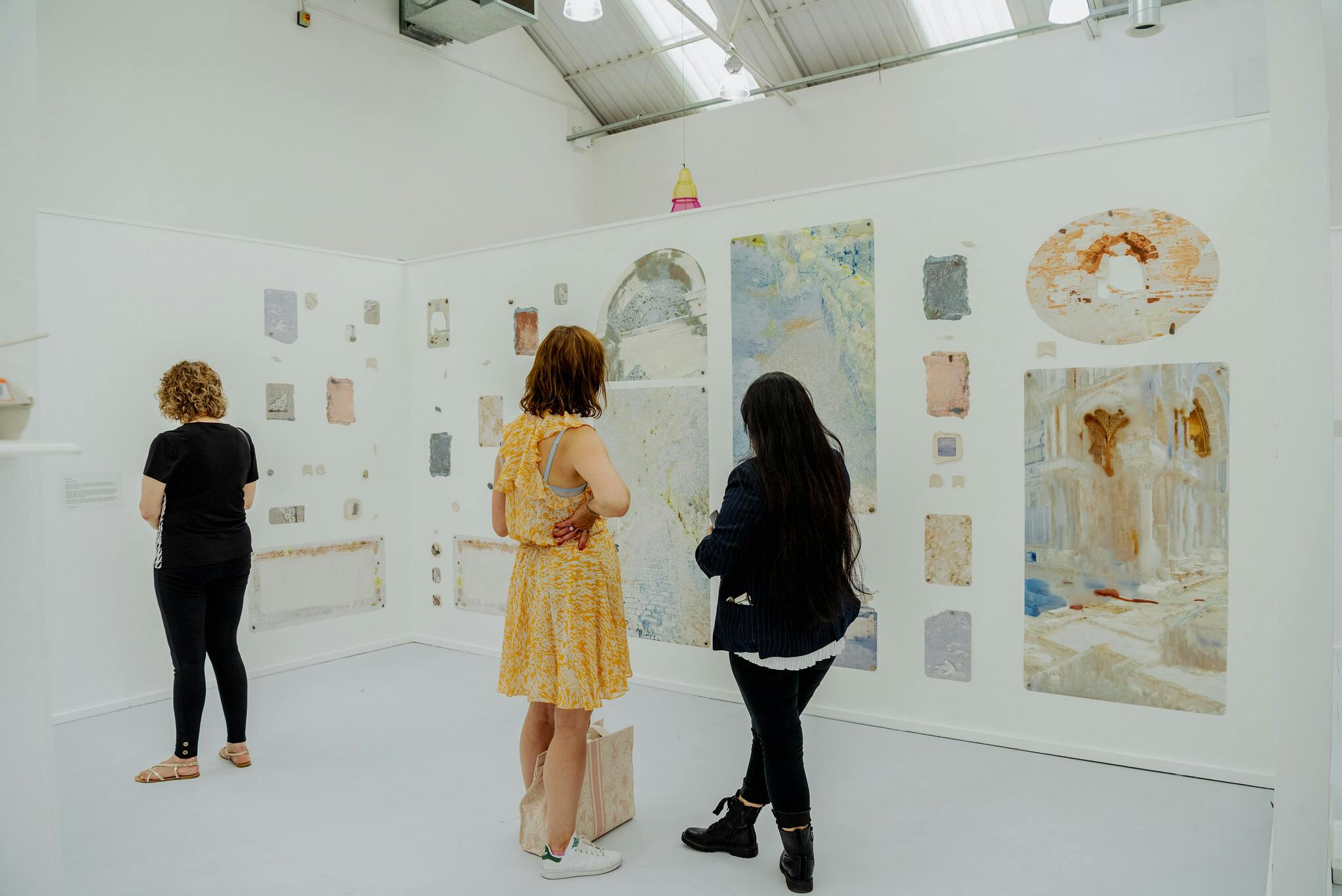 People stand in a light filled gallery with white walls and pastel hued artwork on the walls