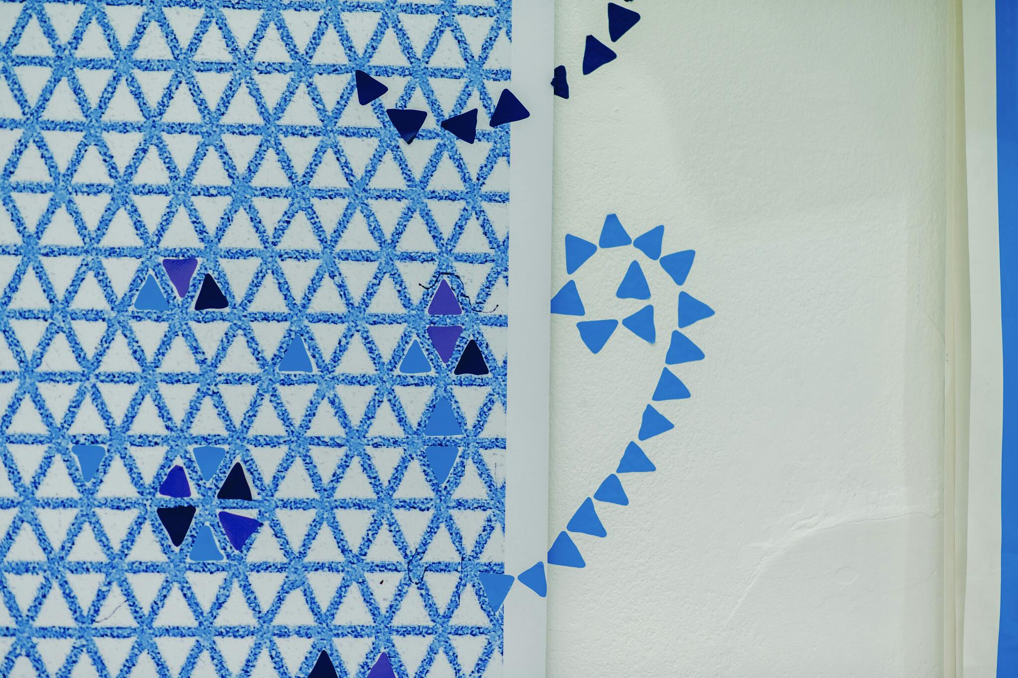 Art work by Tracey Harrison Goldsmith, textured blue triangles adorn a large sheet of paper, some are filled in with the colours black, blue or purple. A spiral of triangles cross from the paper onto the wall.