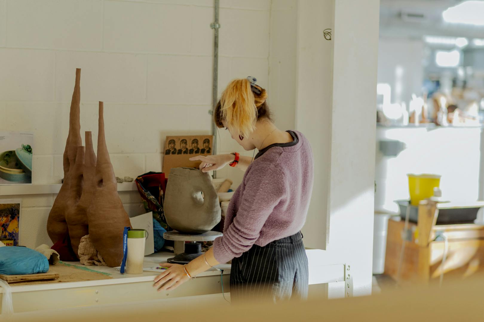 A young woman works on hand building an abstract ceramic vessel at her desk in the dappled light of our ceramics studio. She is surrounded by her other finished ceramic works.