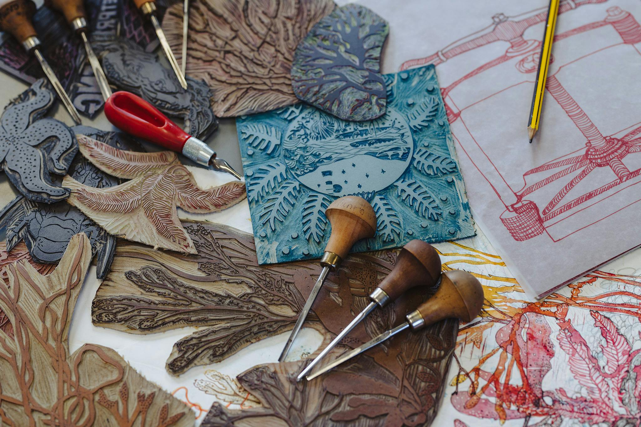 Tools used in lino cutting. Three wooden handles, sharp, pointed chisels lie on carved pieces of lino in the pattern of sea creatures and sea weed. Dommoore 2022 real ideas print in action 72dpi 9829