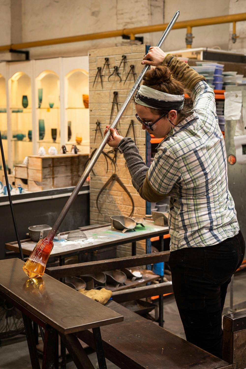 Image shows graduate student Charlotte Scurlock with her hair up in a ponytail, wearing protective goggles and a plaid shirt, manipulating glass in the Teign Valley Glass studio