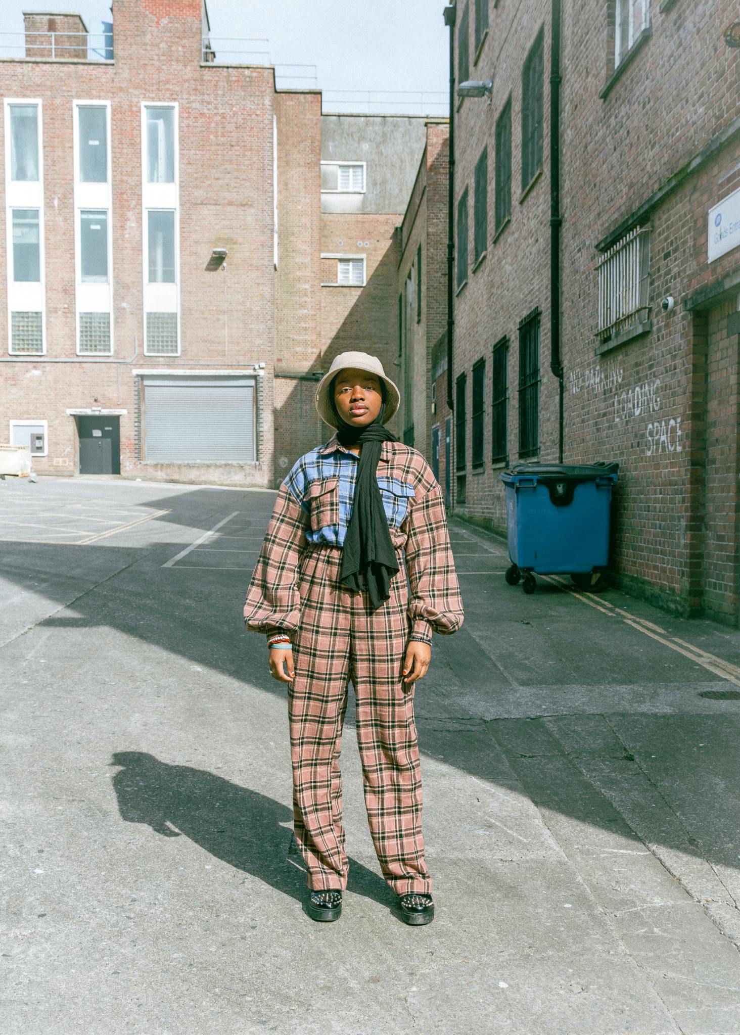 Image shows a woman stood in the middle of a street, with brick buildings around her. She is dressed in a checkered suit and bucket hat and looking directly at the camera.