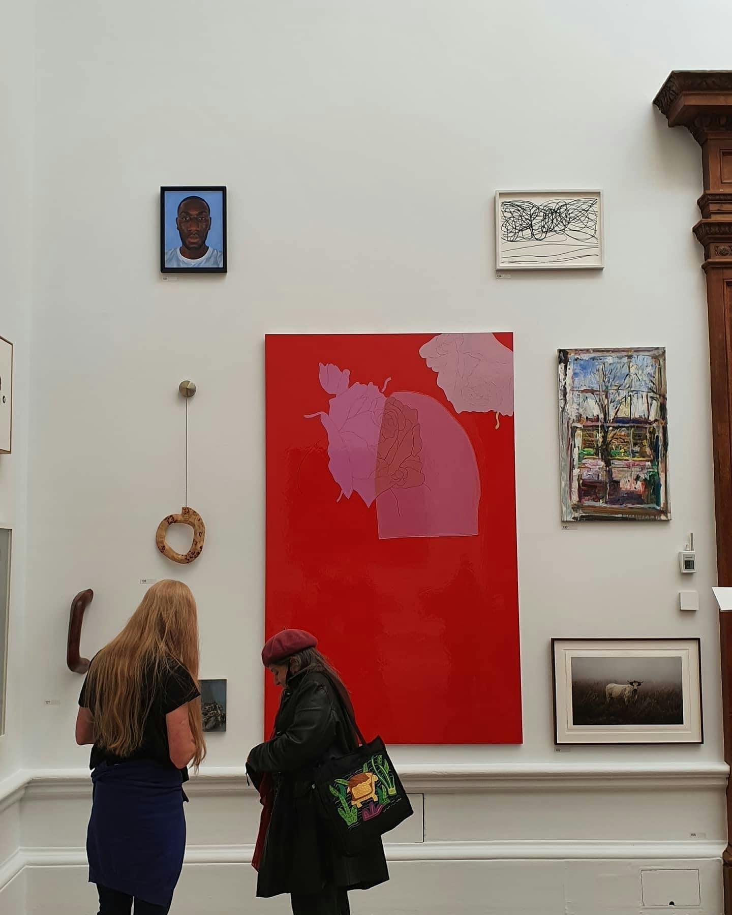Xanthe's painting in place in the Royal Academy of Arts, two people stand to the left hand corner of the photo looking at the paintings, one of which is large and red with pink, surrounded by smaller paintings and framed pictures