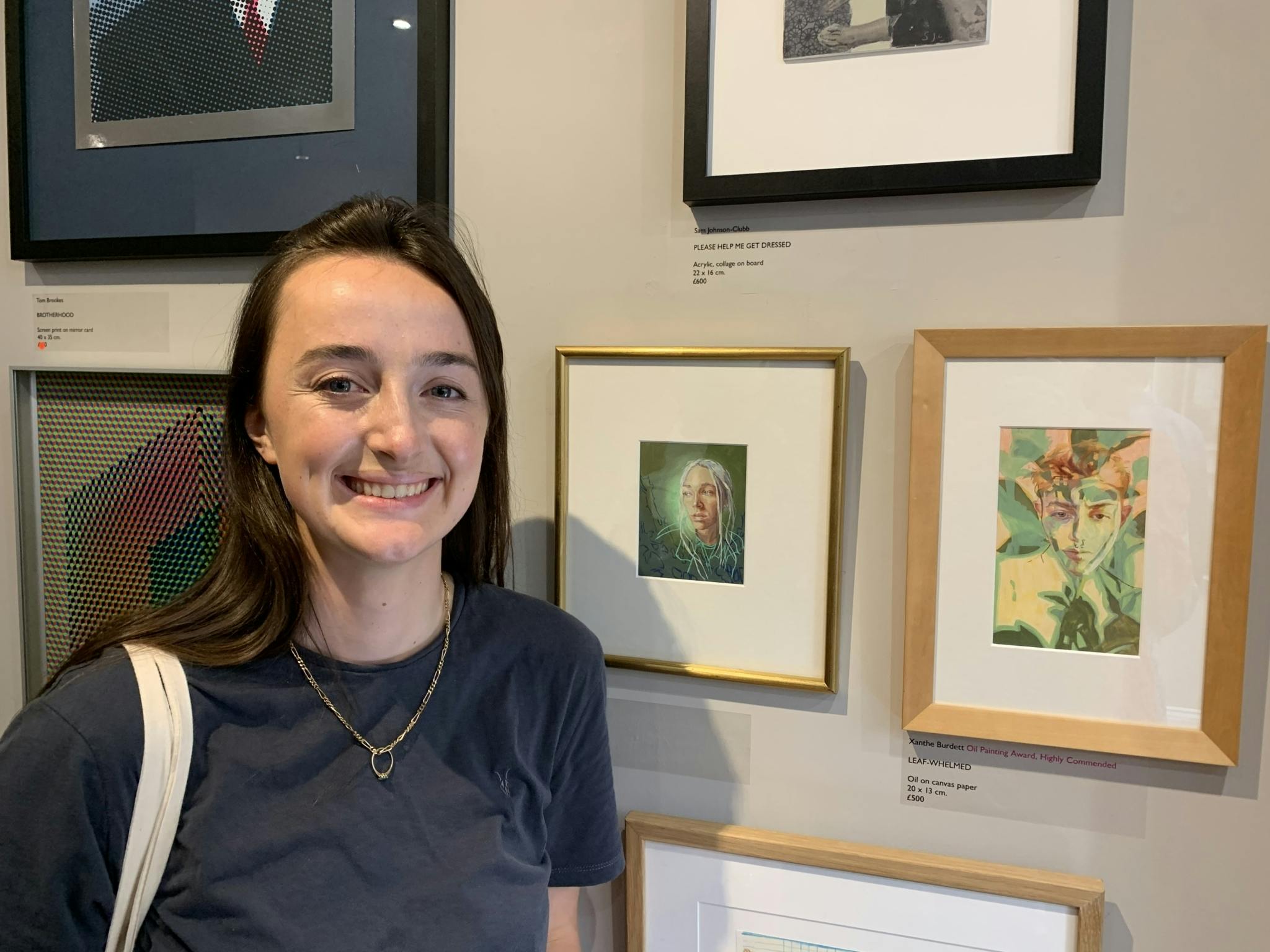 Xanthe, smiling with brown short hair and a grey tshirt, stands in front of a number of small paintings in wooden frames