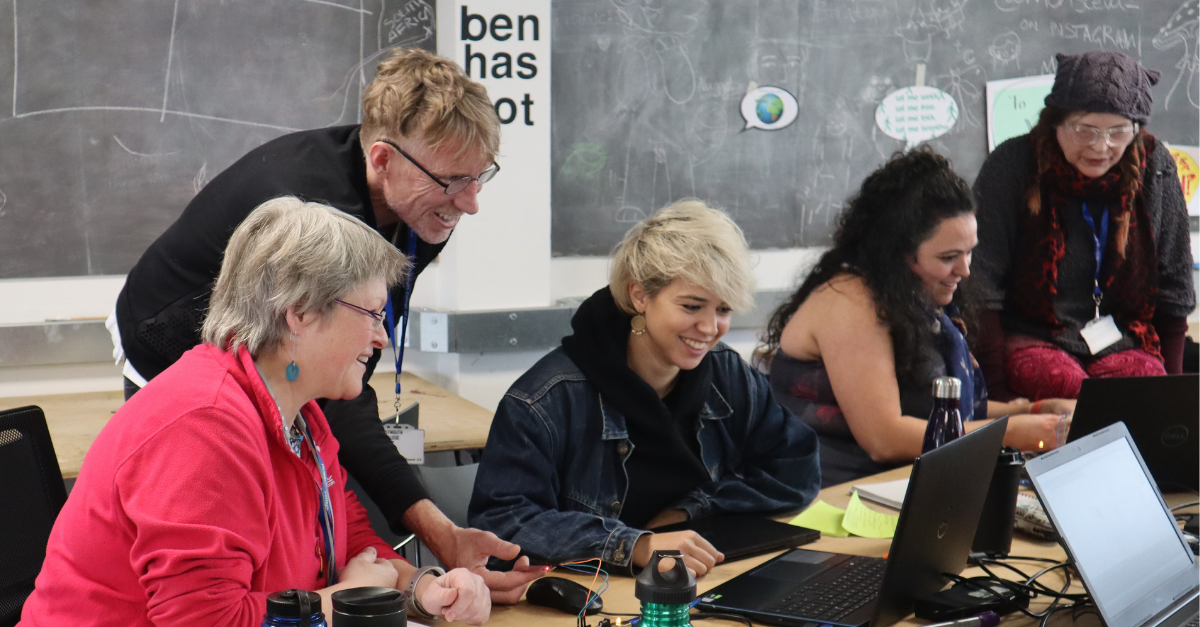 A group of people around a table with laptops and tech materials smile as they learn about technology during a Fab Lab Tech and Business Bootcamp