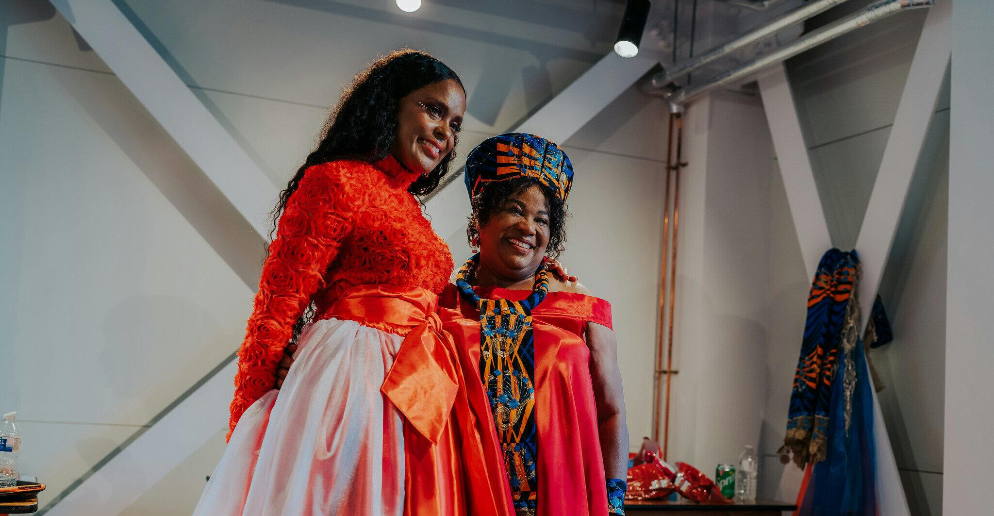 Two women smiling backstage at a fashion show, one of them is wearing a pink red top made up of fabric roses and a pink long skirt with an orange bow around the waist. The next is wearing a blue & orange hat and dress inspired by her African heritage.