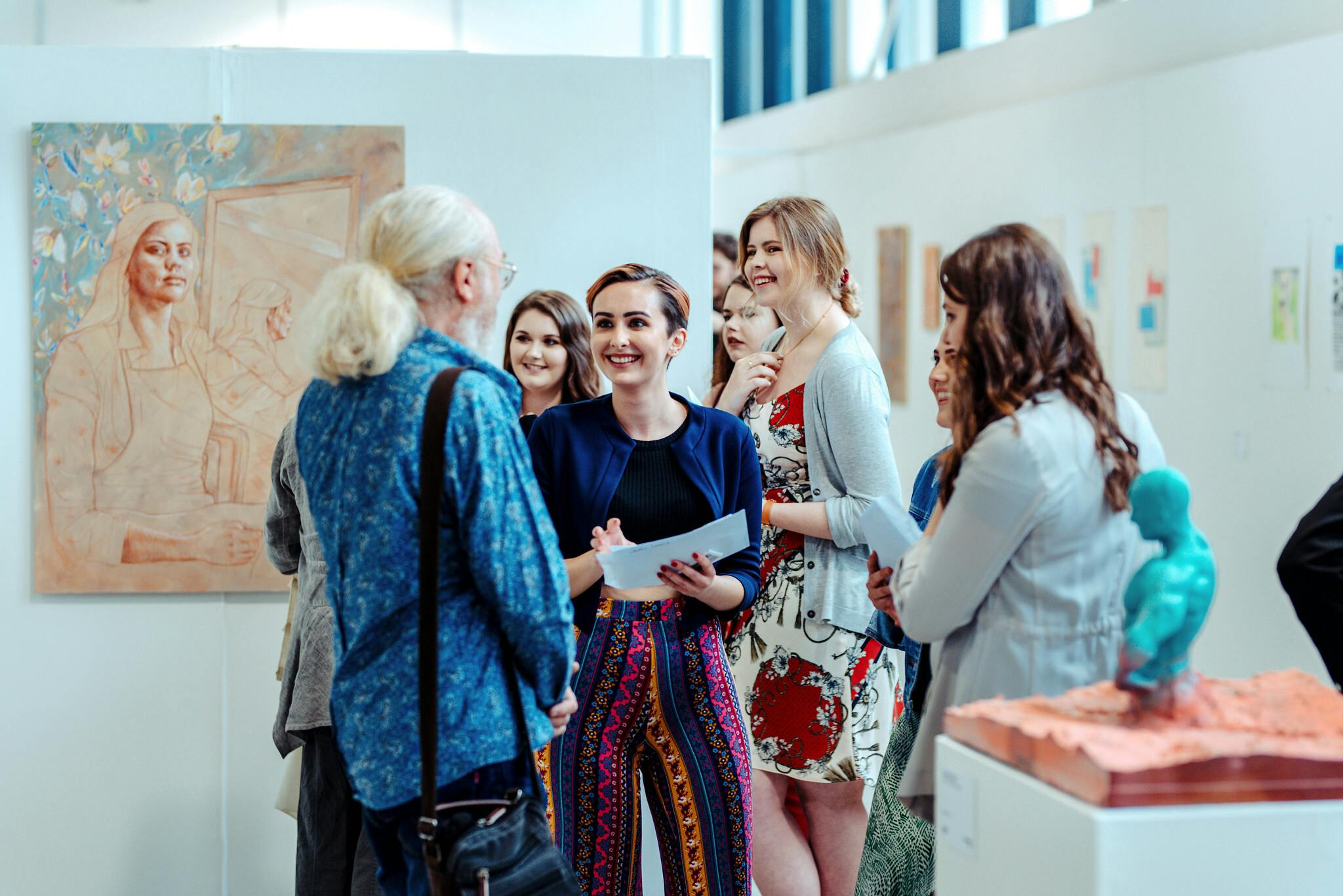 Four women smile and speak to a man in a gallery space with paintings hung on the walls
