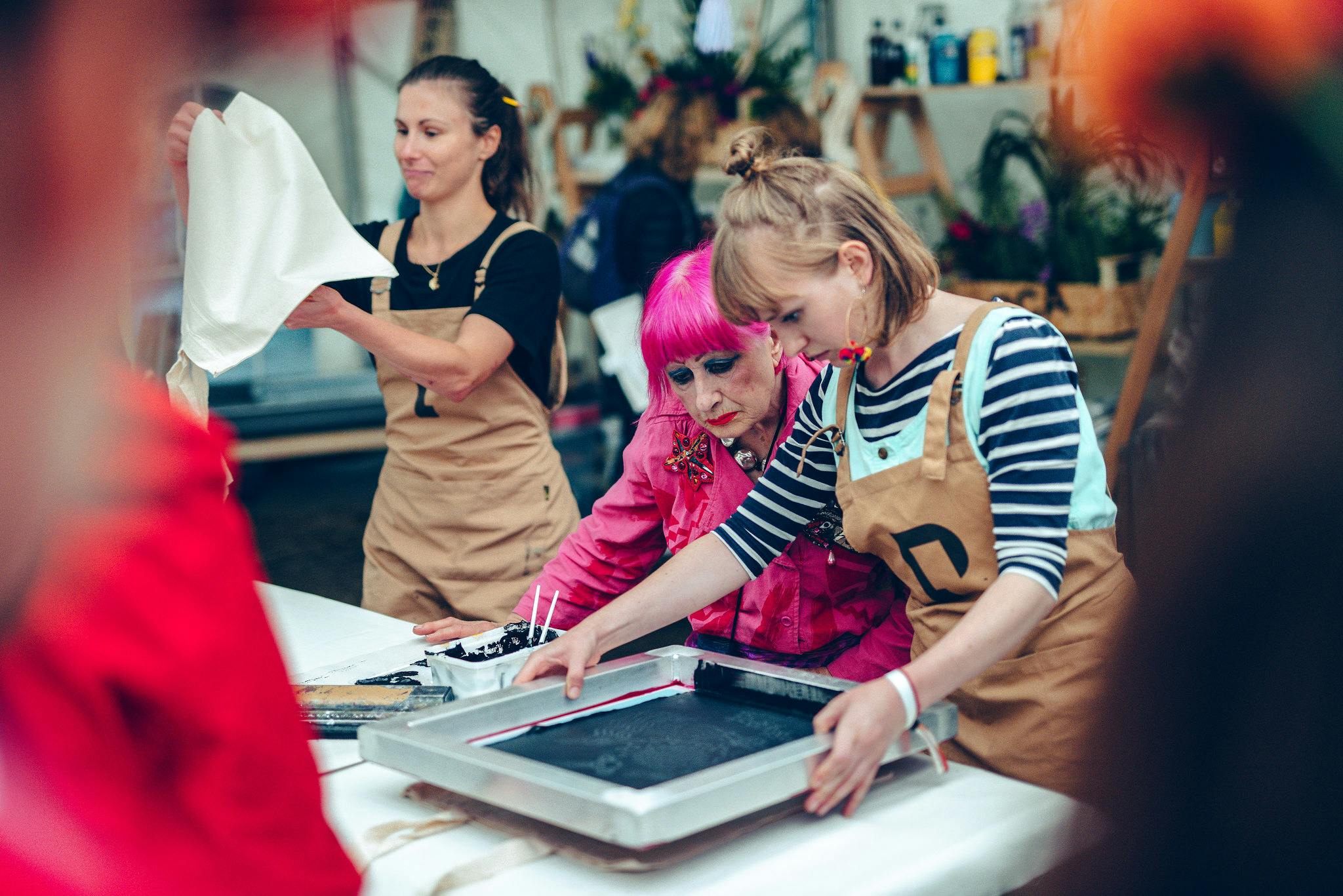 Fashion and textile designer Zhandra Rhodes sporting hot pink hair and a matching pink boilersuit leads a screenprinting demonstration within a busy festiva tent, surrounded by plants and art supplies.