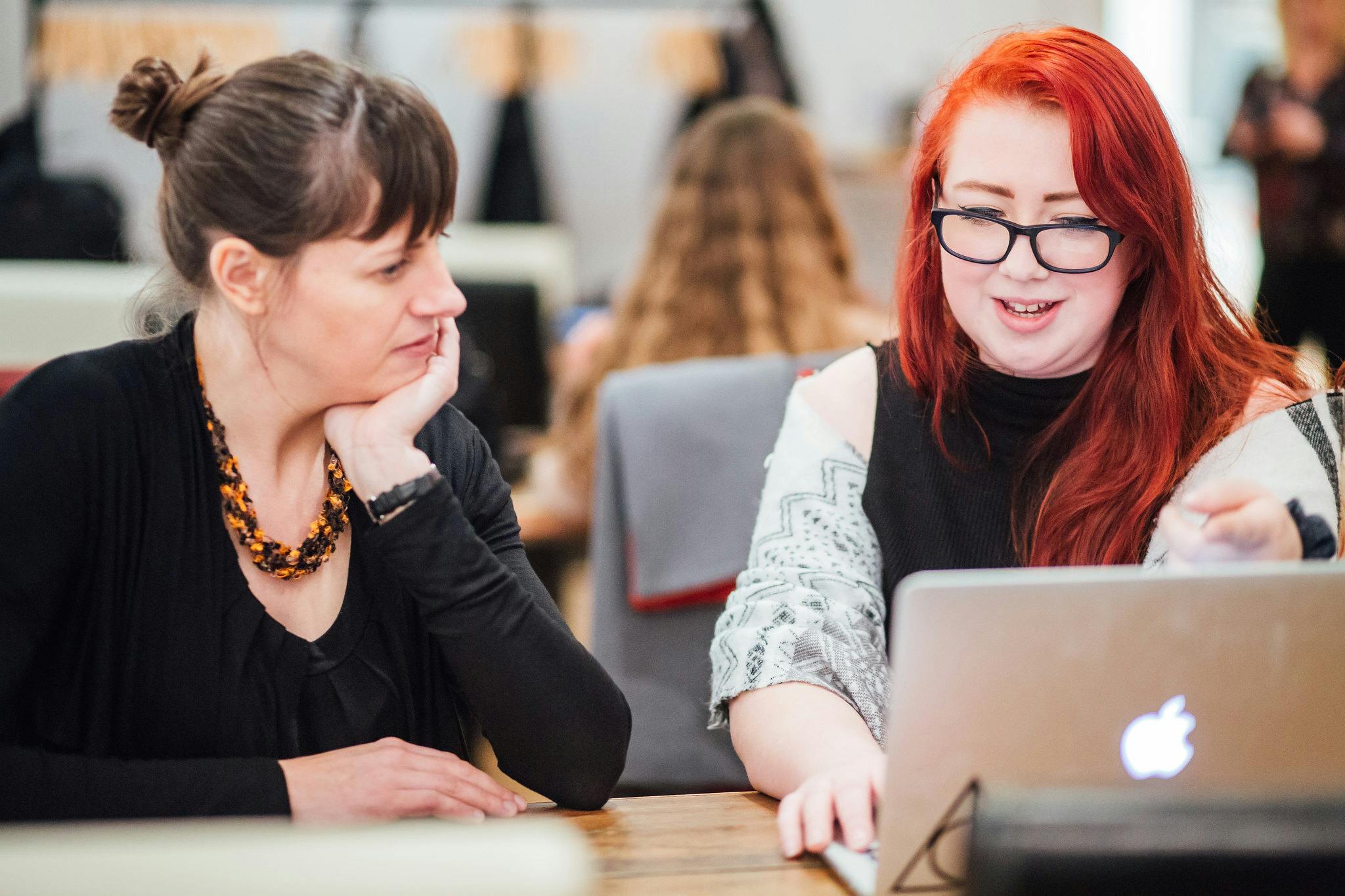 A young woman with red hair and glasses shows her portfolio on her laptop to an female industry photographer in a one to one portfolio review.