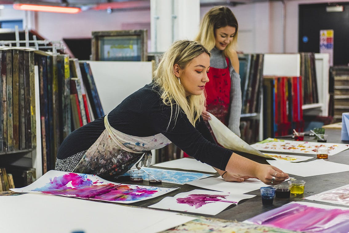 A student leans across a print table covered in large sheets of paper coloured with inks ready for heat transfer printing to fabric.