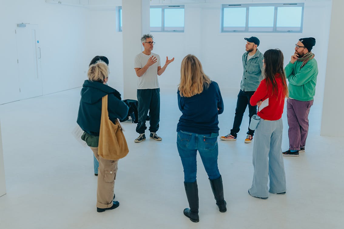 Postgraduate students stand in a white walled room listening to artist Oliver Frank Chanarin speaking at Karst gallery in Plymouth