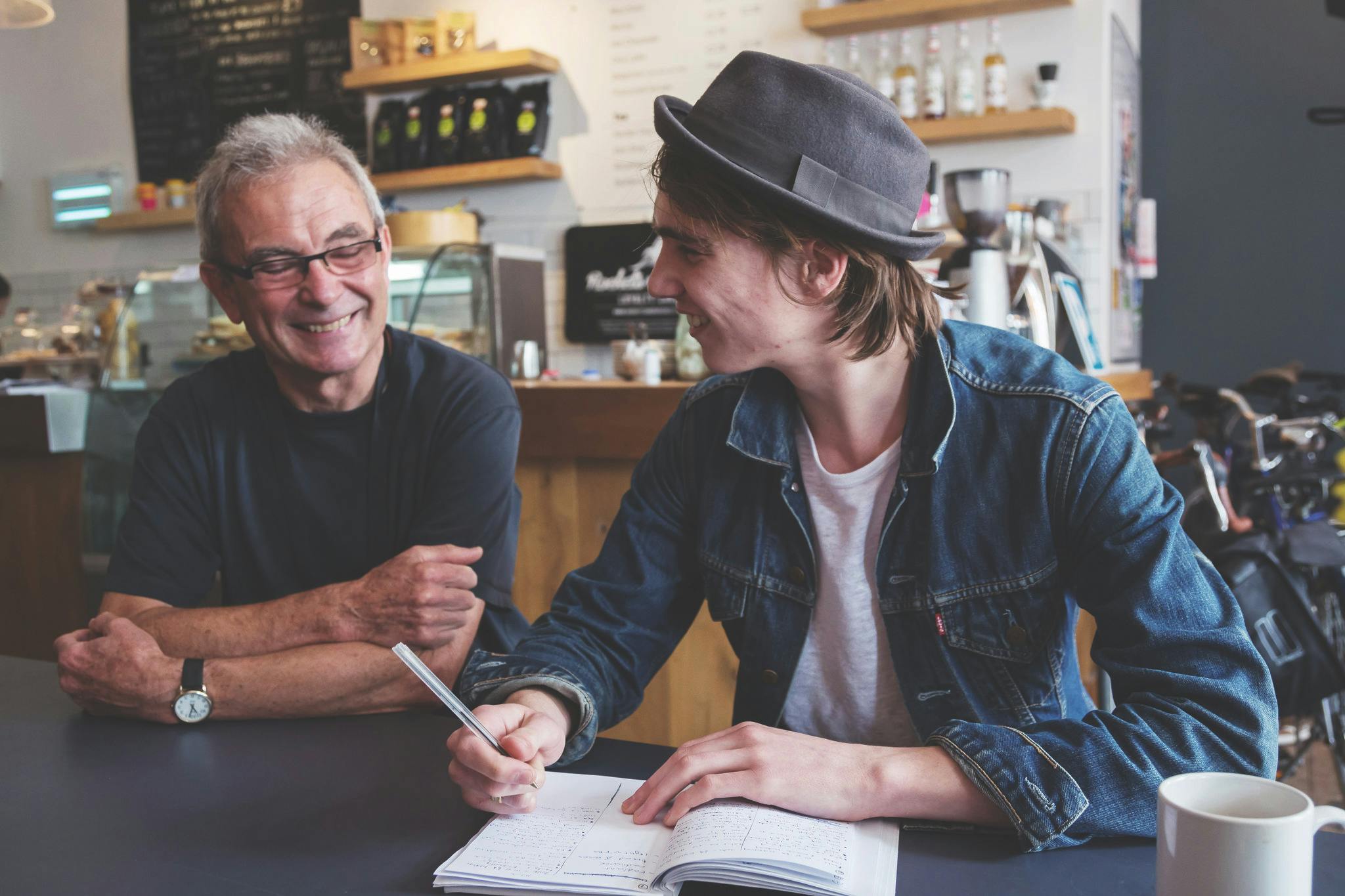 Two people, one young and the other older, sit in a cafe smiling and writing in a notebook