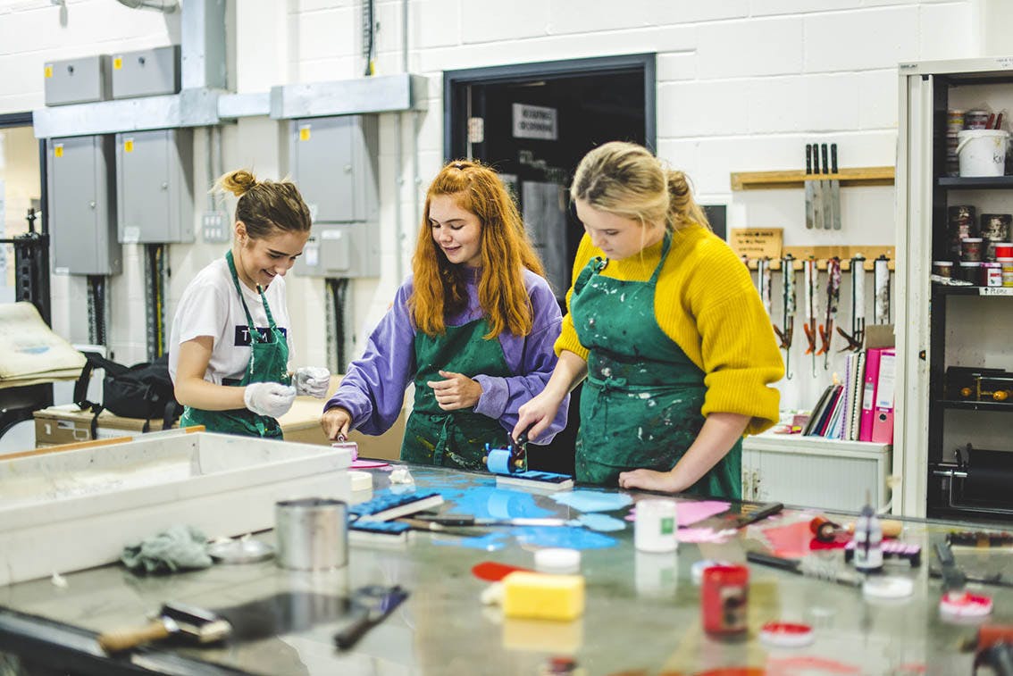 Gathered around a printing station, three female students laugh in paint-splatted aprons whilst hand-rolling ink.