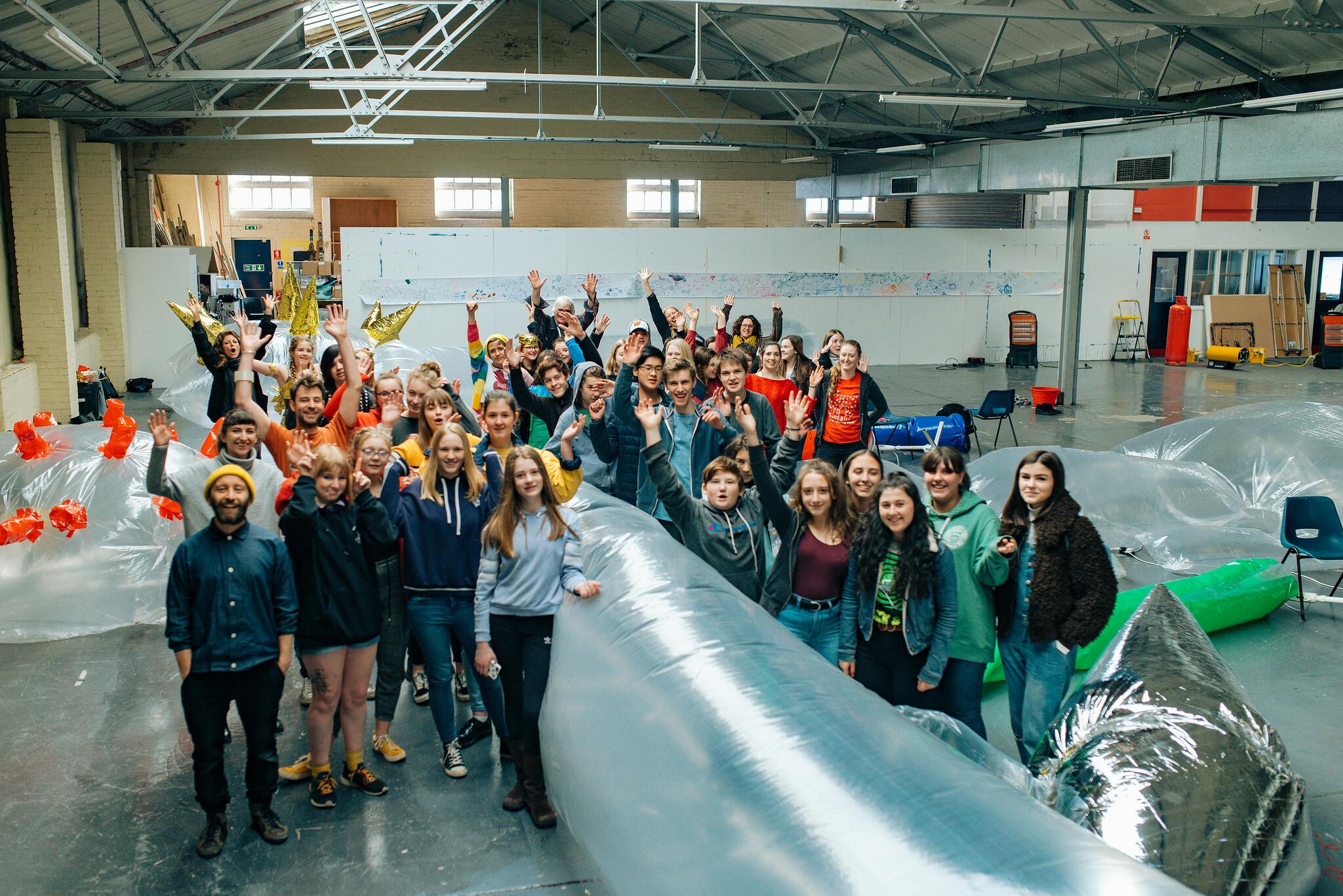 A large class of school-age students pose for a photo in an industrial-looking warehouse with inflatable installations.