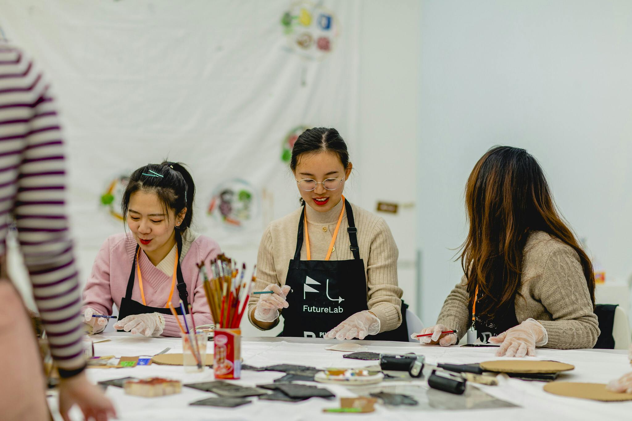 Three girls sit at a table in black aprons, on the table are lino stencils and blocks which they are rolling ink onto and printing with.