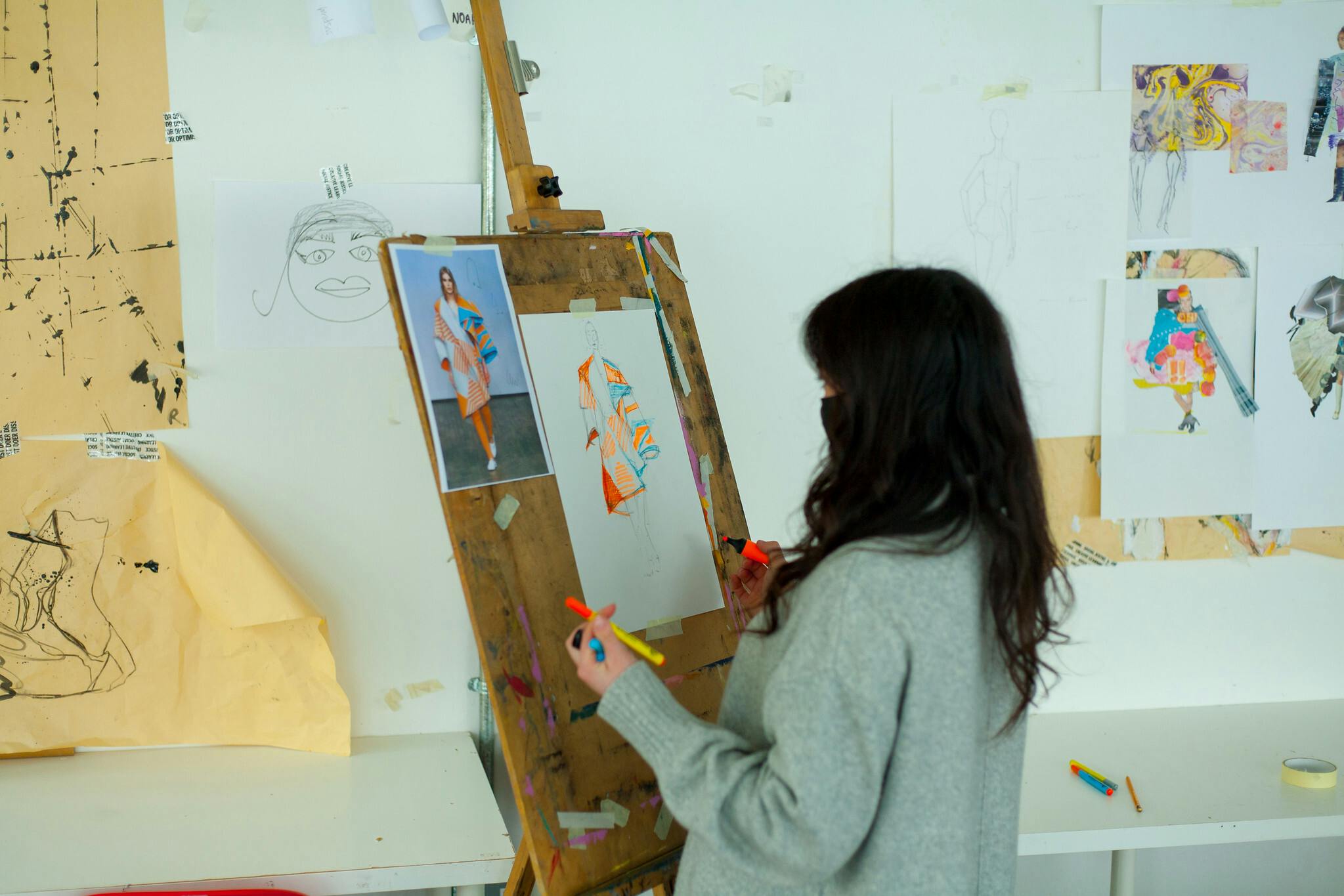 A girl stands at an easel against a wall covered in paintings and sketches. The easel has paper pinned onto it with the beginnings of a fashion illustration