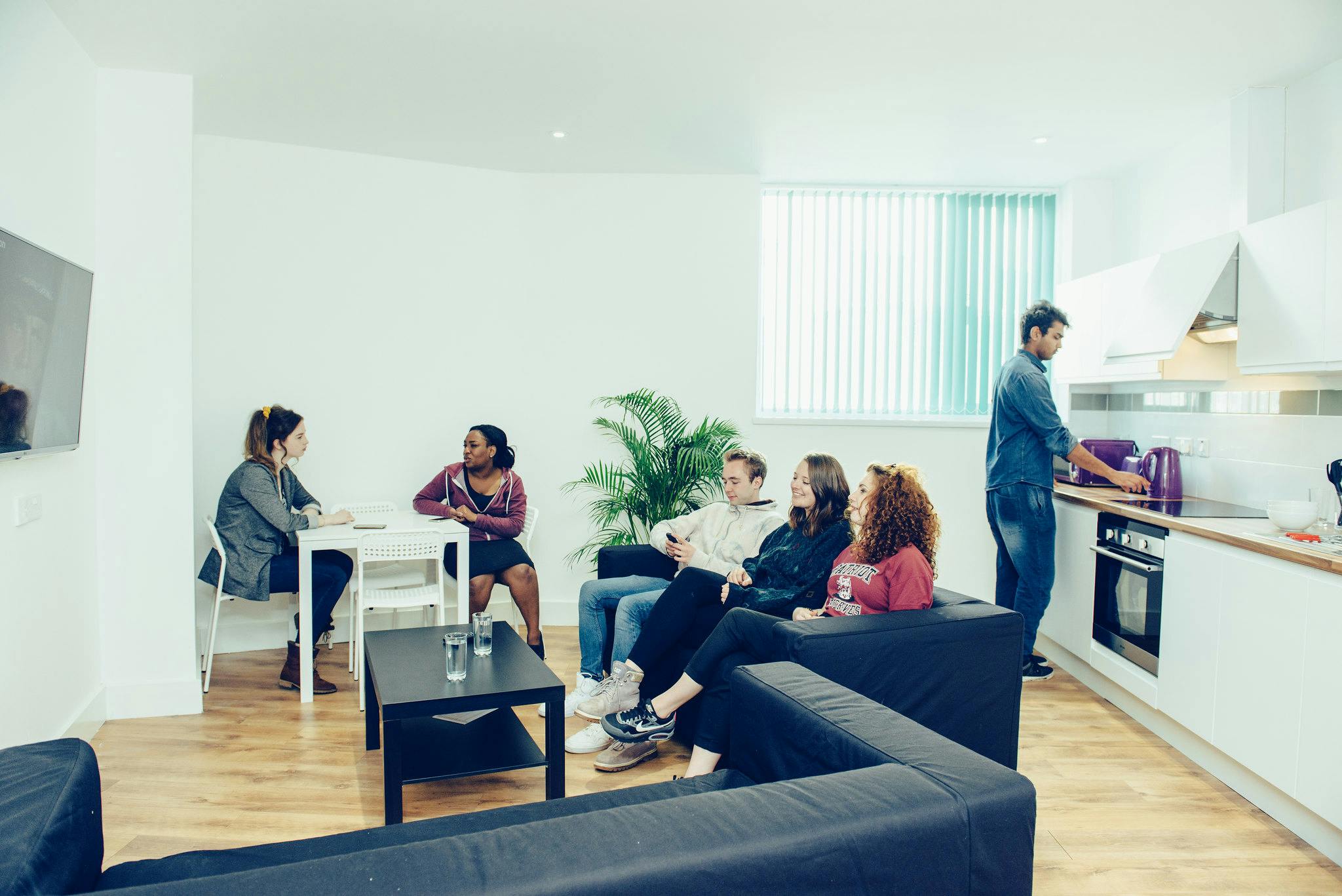 Five students sit on a black sofa, at a white table, whilst one puts the kettle on in the kitchen. The space is bright and clean and is one of Clever Student Lets' shared halls.