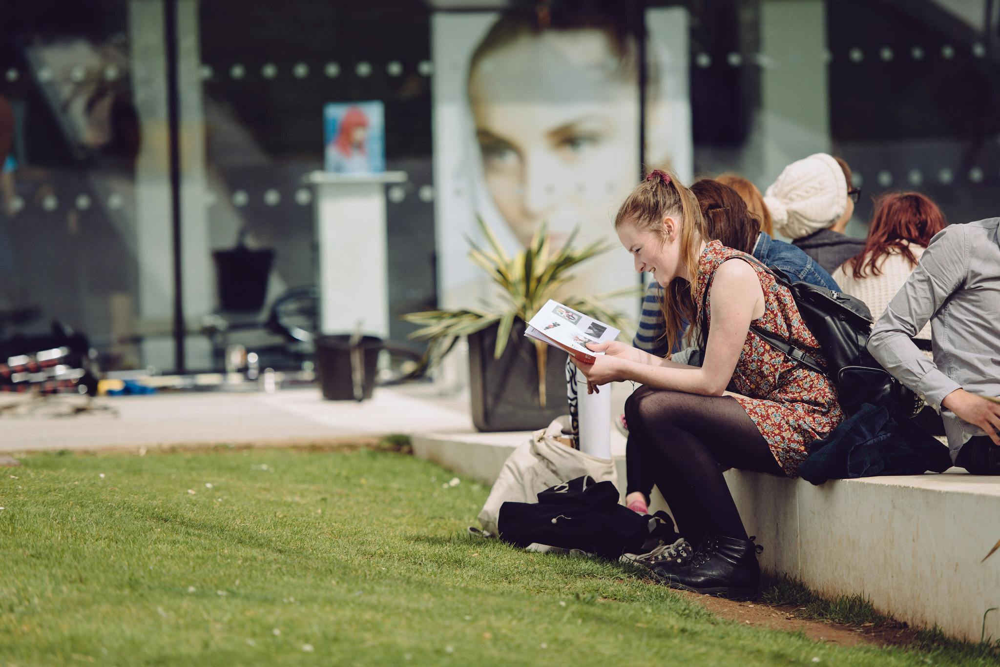 "A female student sits in a grassy area with fellow students on a concrete bench outside The Gallery window smiling whilst reading"