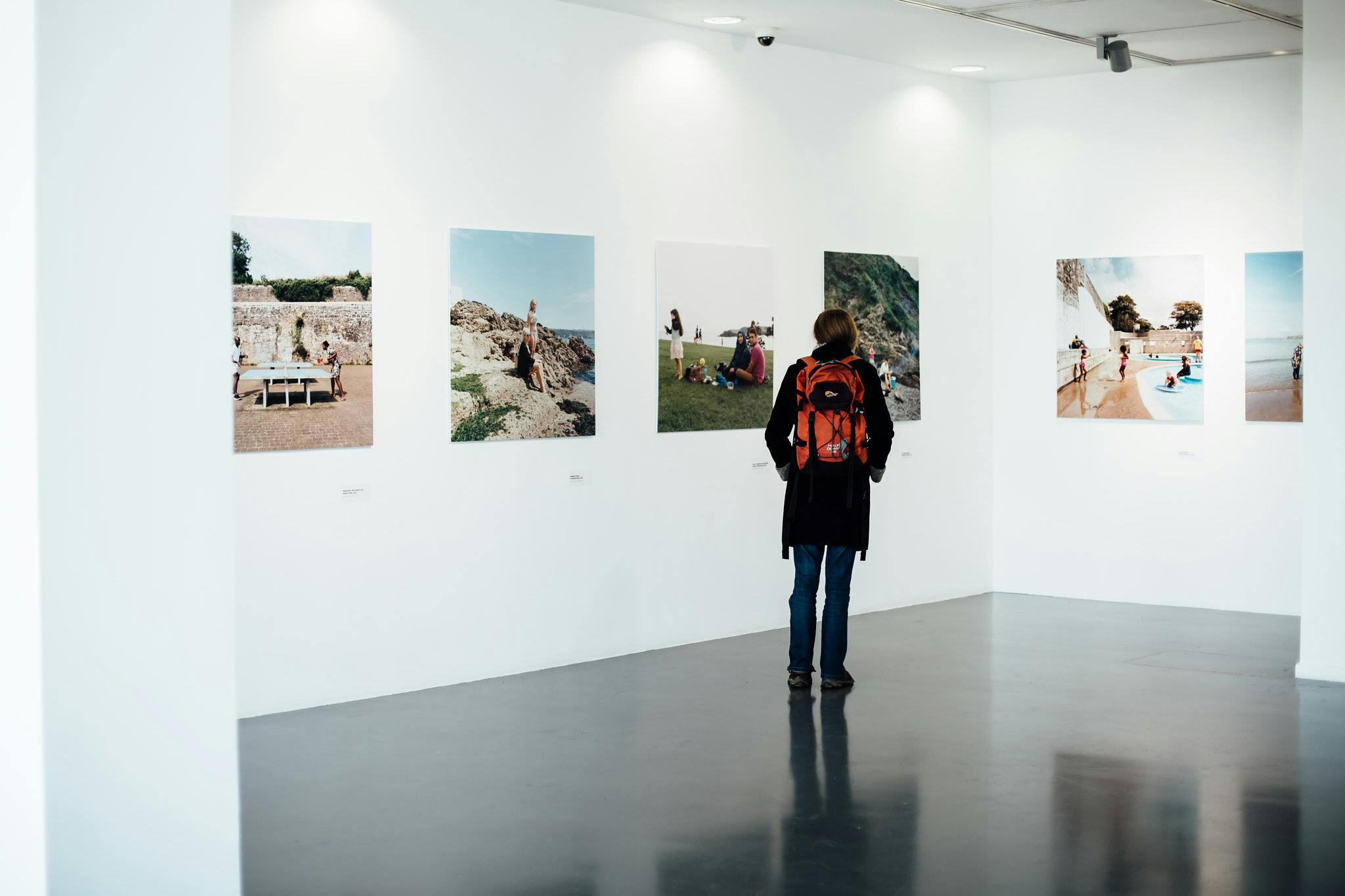 A woman stands in a white gallery space looking at large printed photographs of people at the seaside.