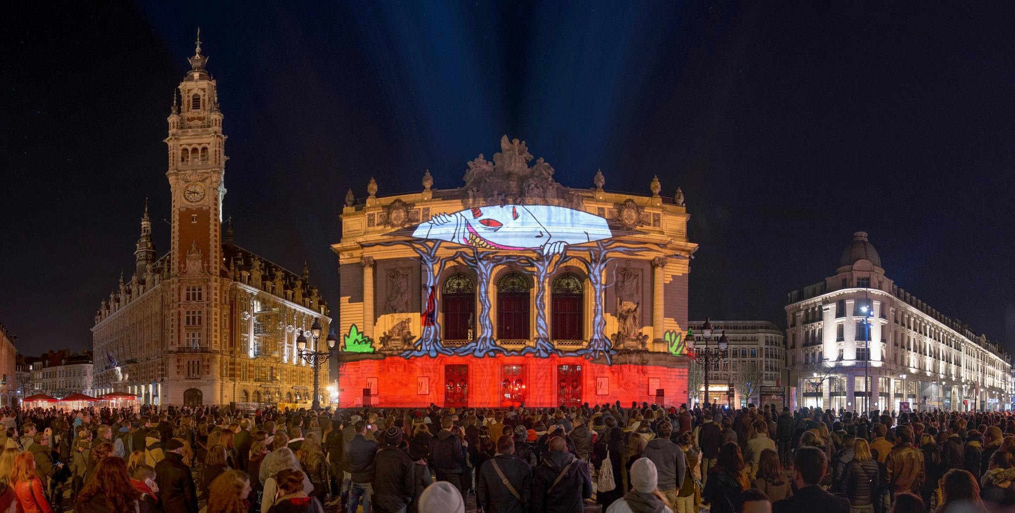 An evening in the city of Lille, France looking out onto a large crowd all watching a videomapped projection on an old building.