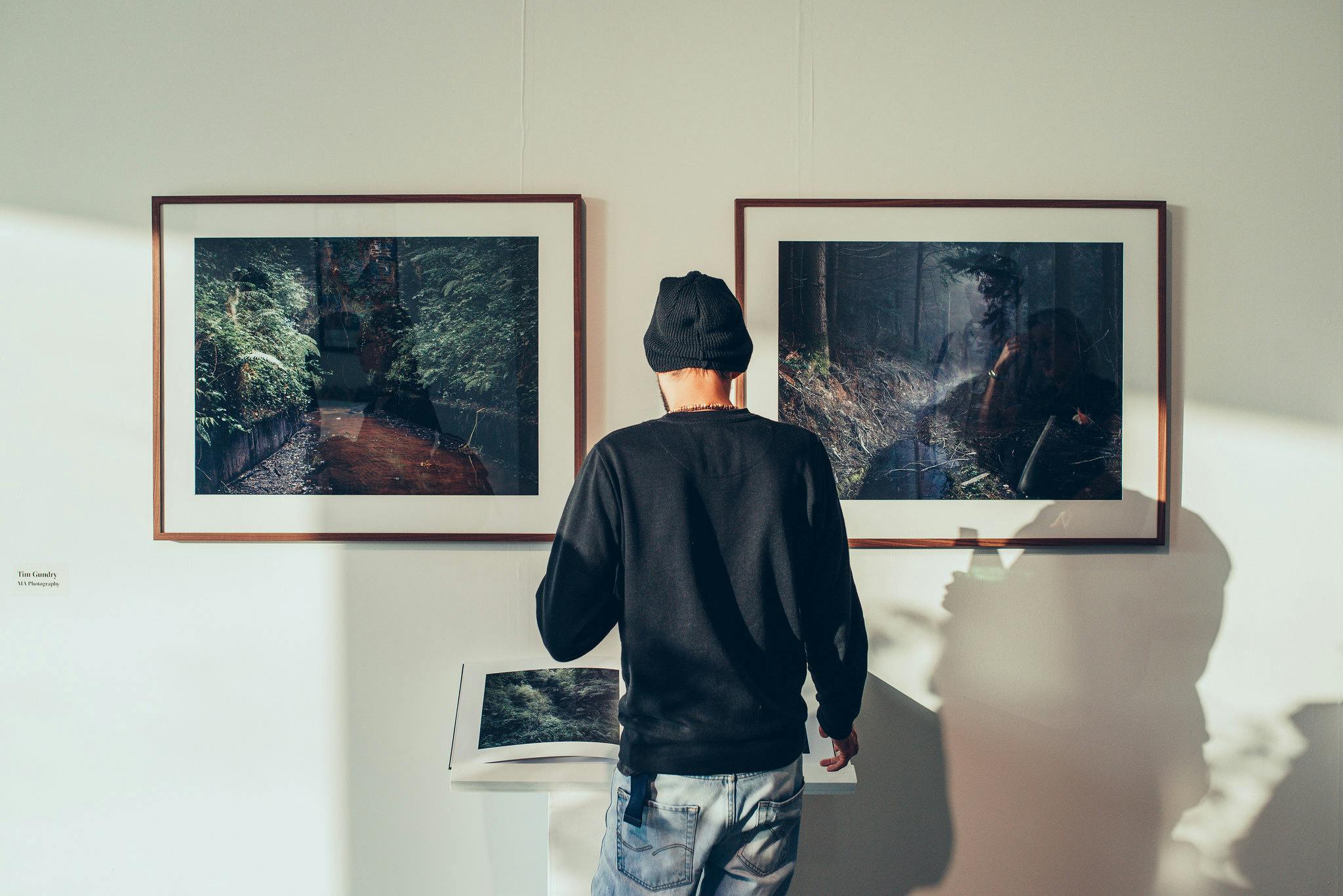 A man stands in a white gallery space looking at two framed photographs showing landscapes of forests.