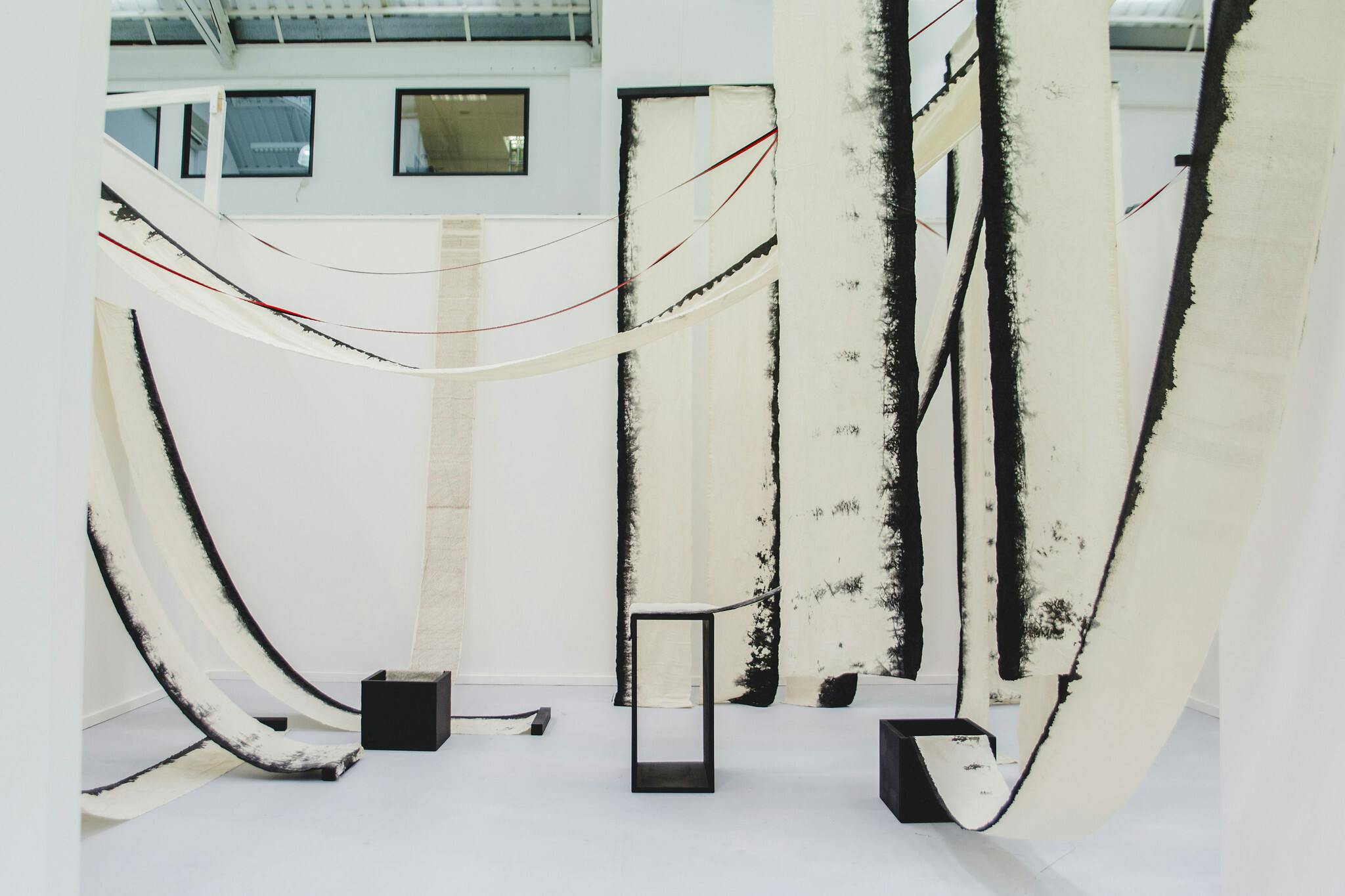 In a white exhibition space, long pieces of off-white fabric hang from the ceiling with the ends tucked into black boxes. The material has black edges with small text printed on it.