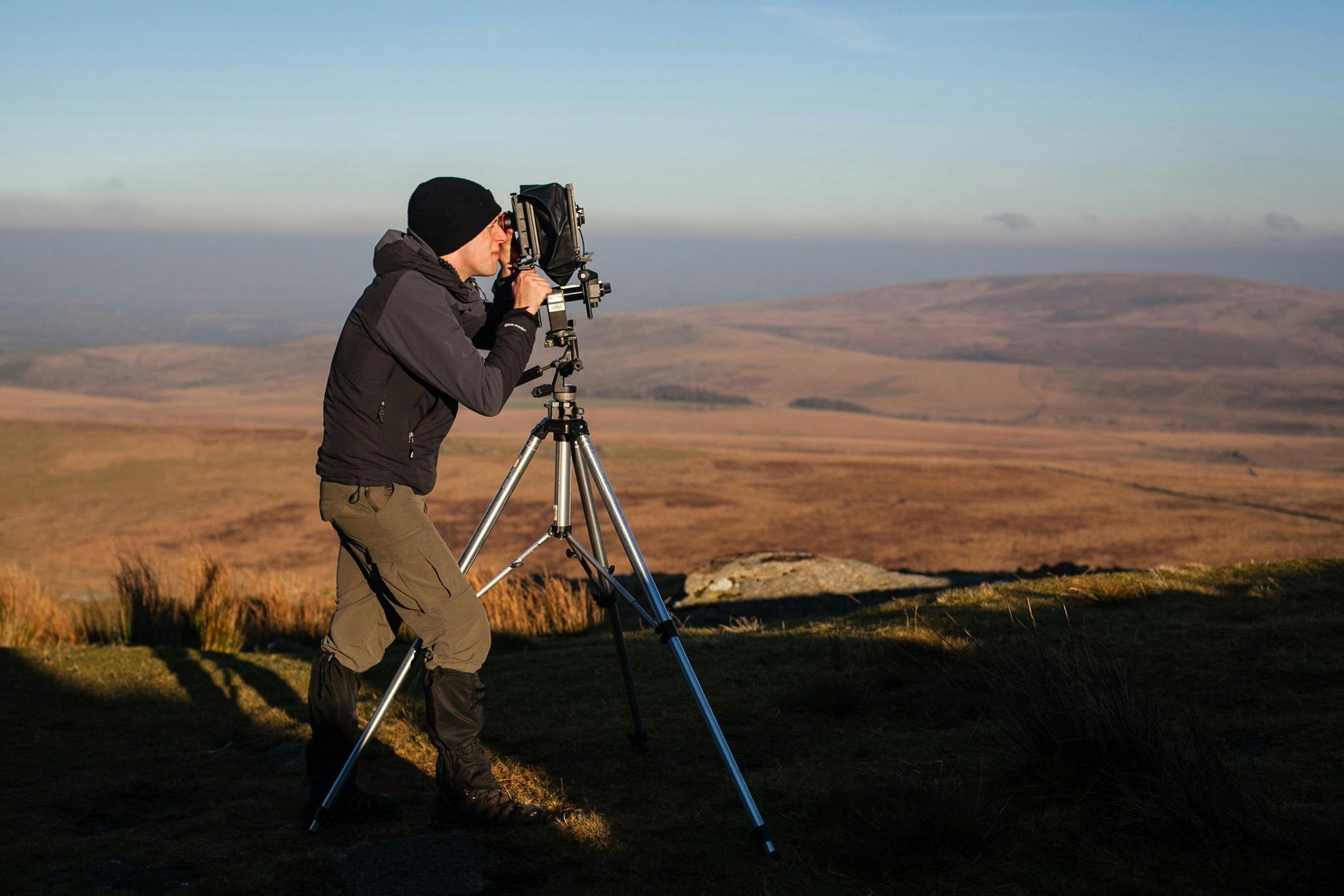 Graduate Nicholas White stands at the top of a tor in Dartmoor looking through the lens of a large 5x4 film camera as the rich golden light of the morning sun brings the moorland to life around him.