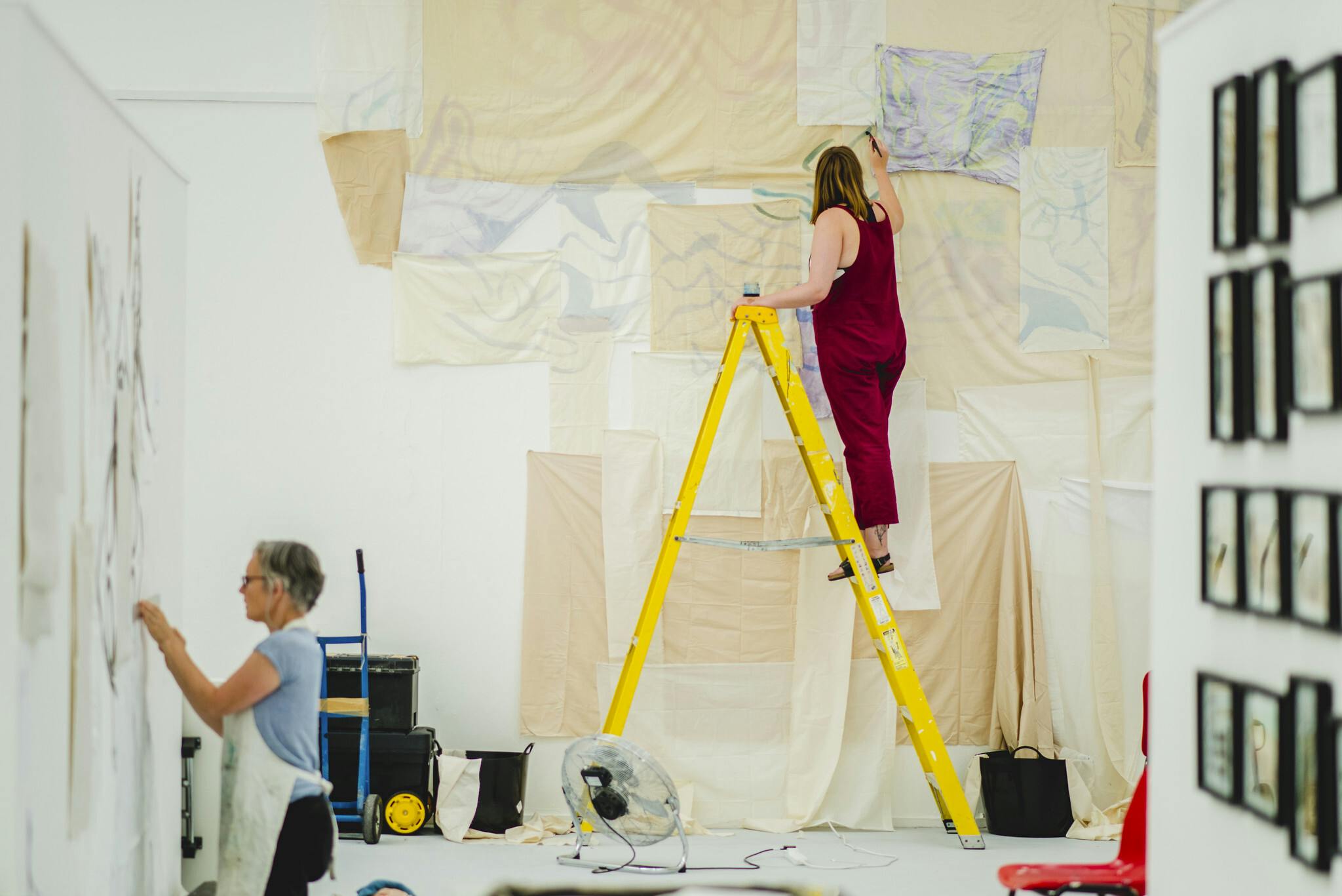 Two female students take over the postgraduate studios, one up a bright yellow ladder layering neutral shades of cloth and paper, the other kneeling on the floor to carefully pin her work.