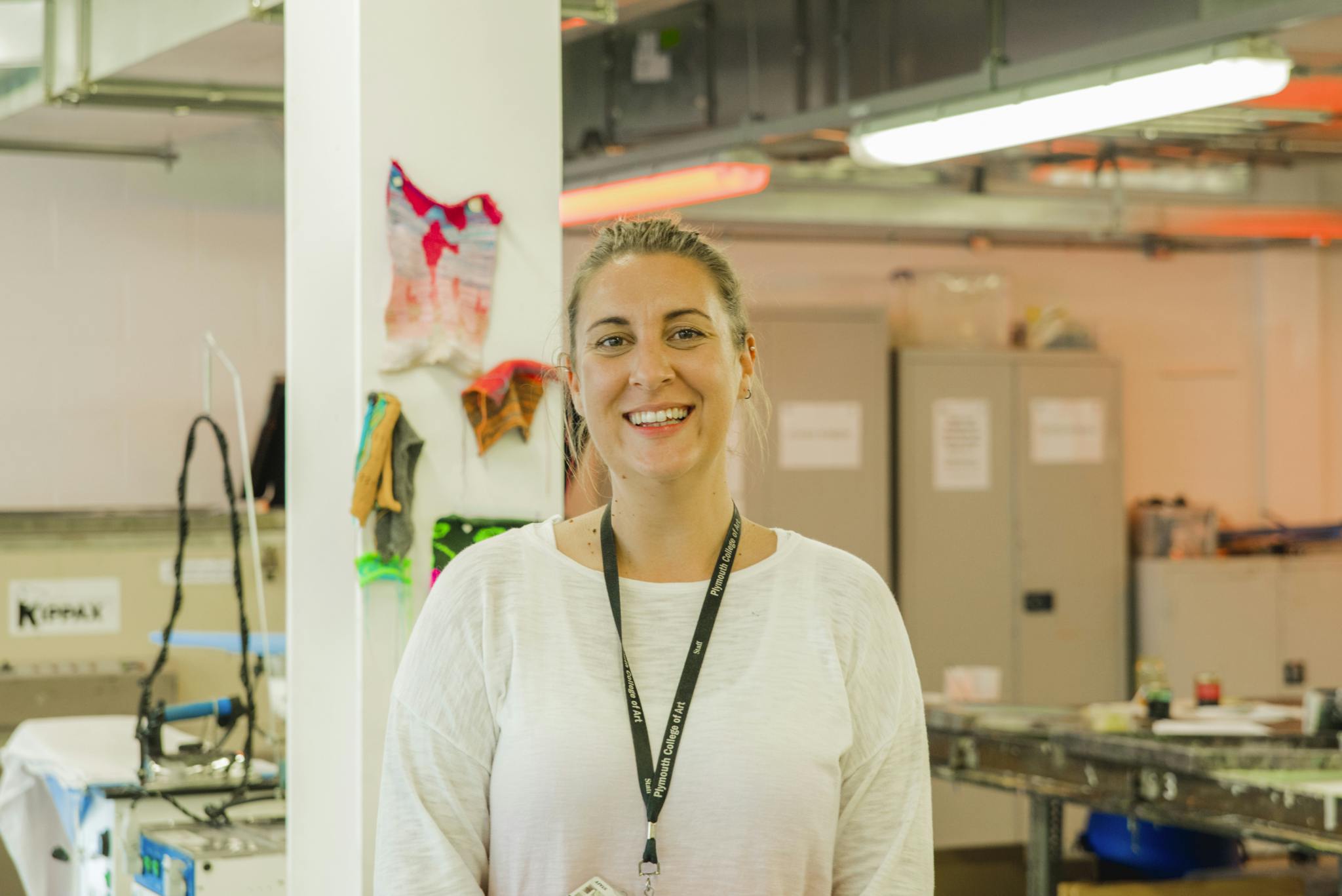 Kathryn Hays wearing a white top and her Plymouth College of Art lanyard stands smiling and relaxed in front of the Fabric Lab, a textiles resource and facility for students