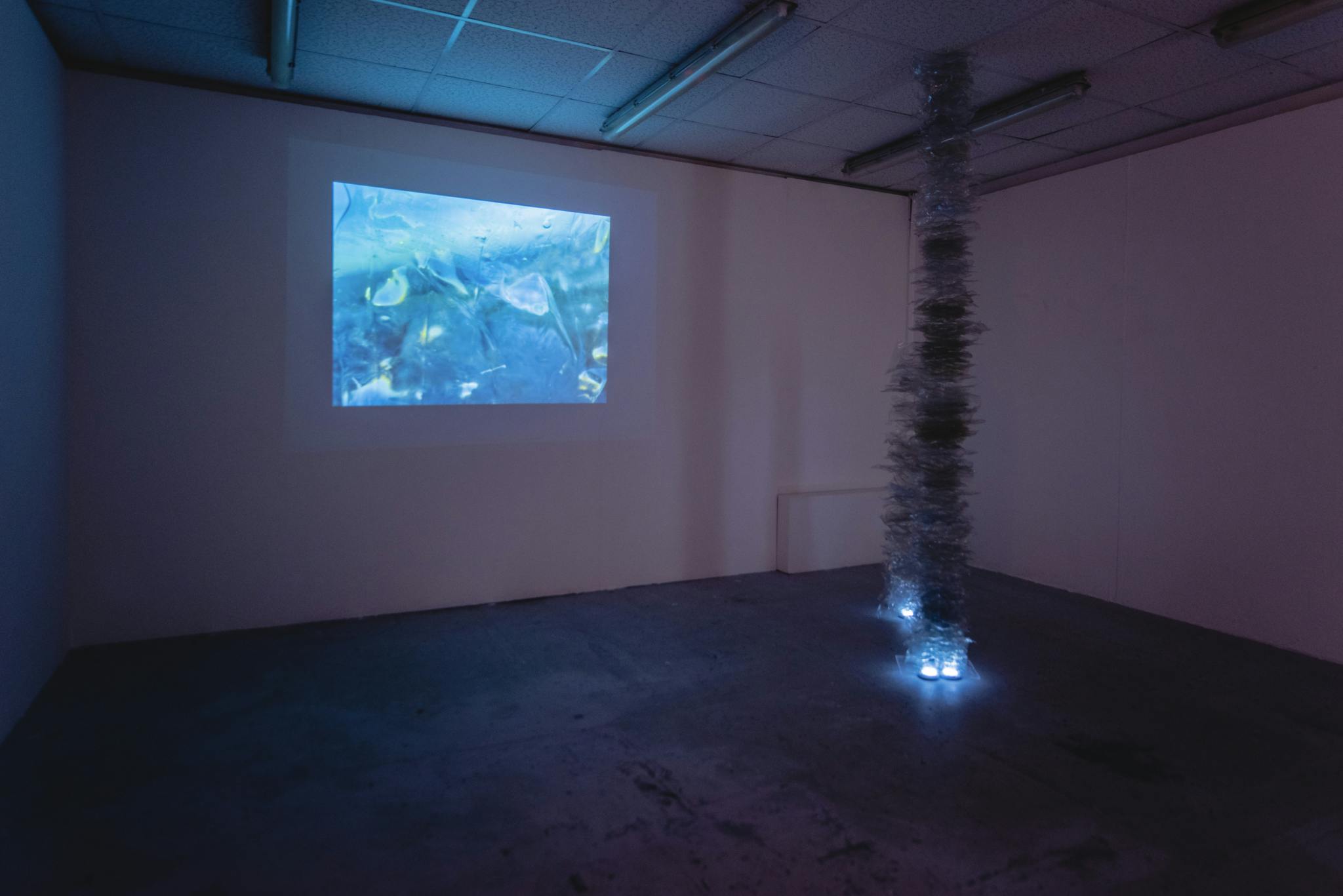Image shows an art installation featuring a pole covered in shredded plastic being lit up from the bottom with a projected film in the background on the wall