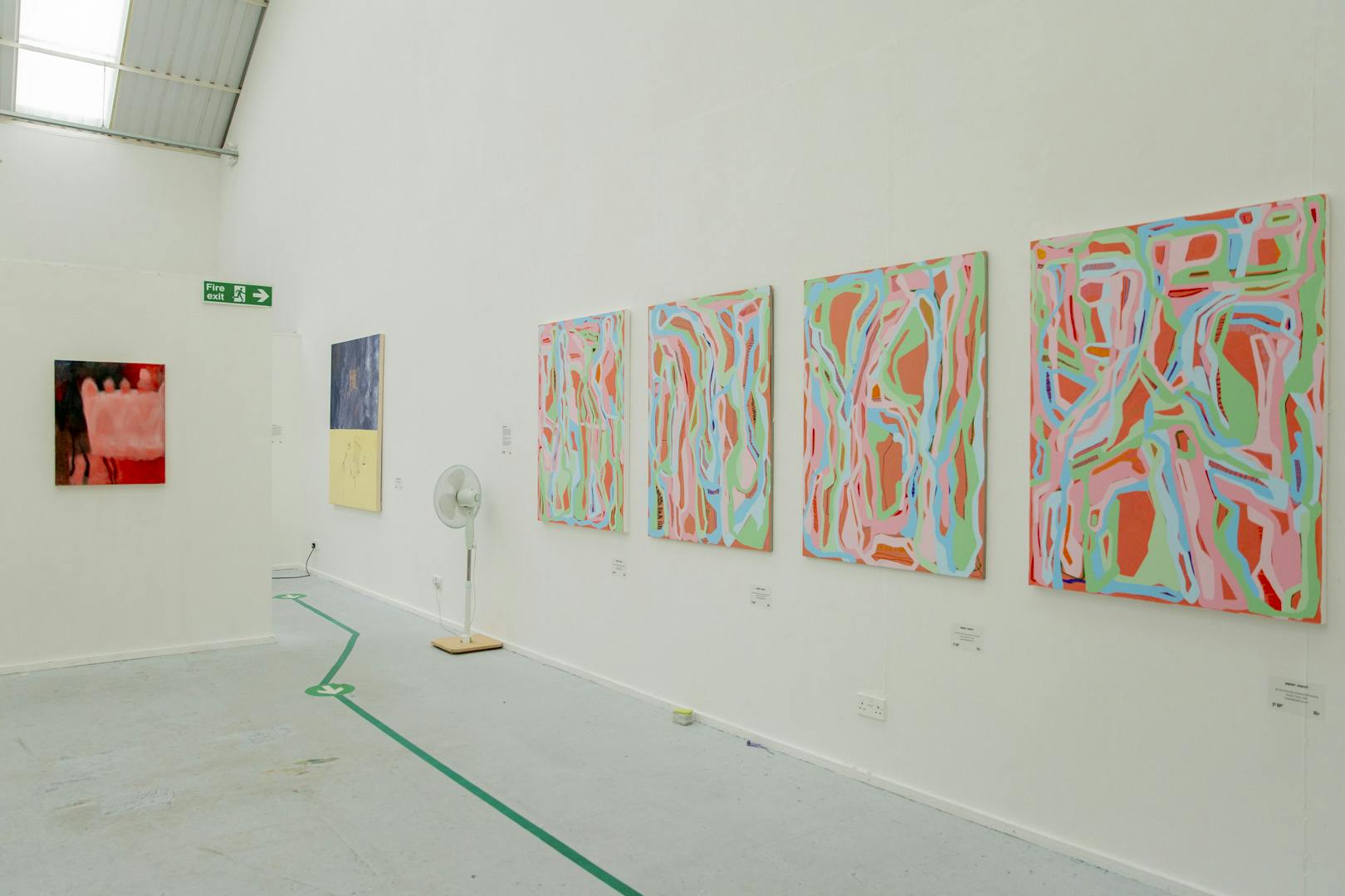 Image shows Ebony's four paintings in situ in the exhibition, featuring bright colourful patterns against a white neutral background with other student work in the background