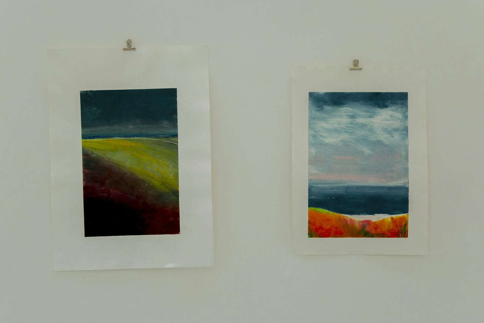 An image of two of Martine's paintings that are on paper and hung by bulldog clips, the paintings show landscape imagery with interesting colour palette choices