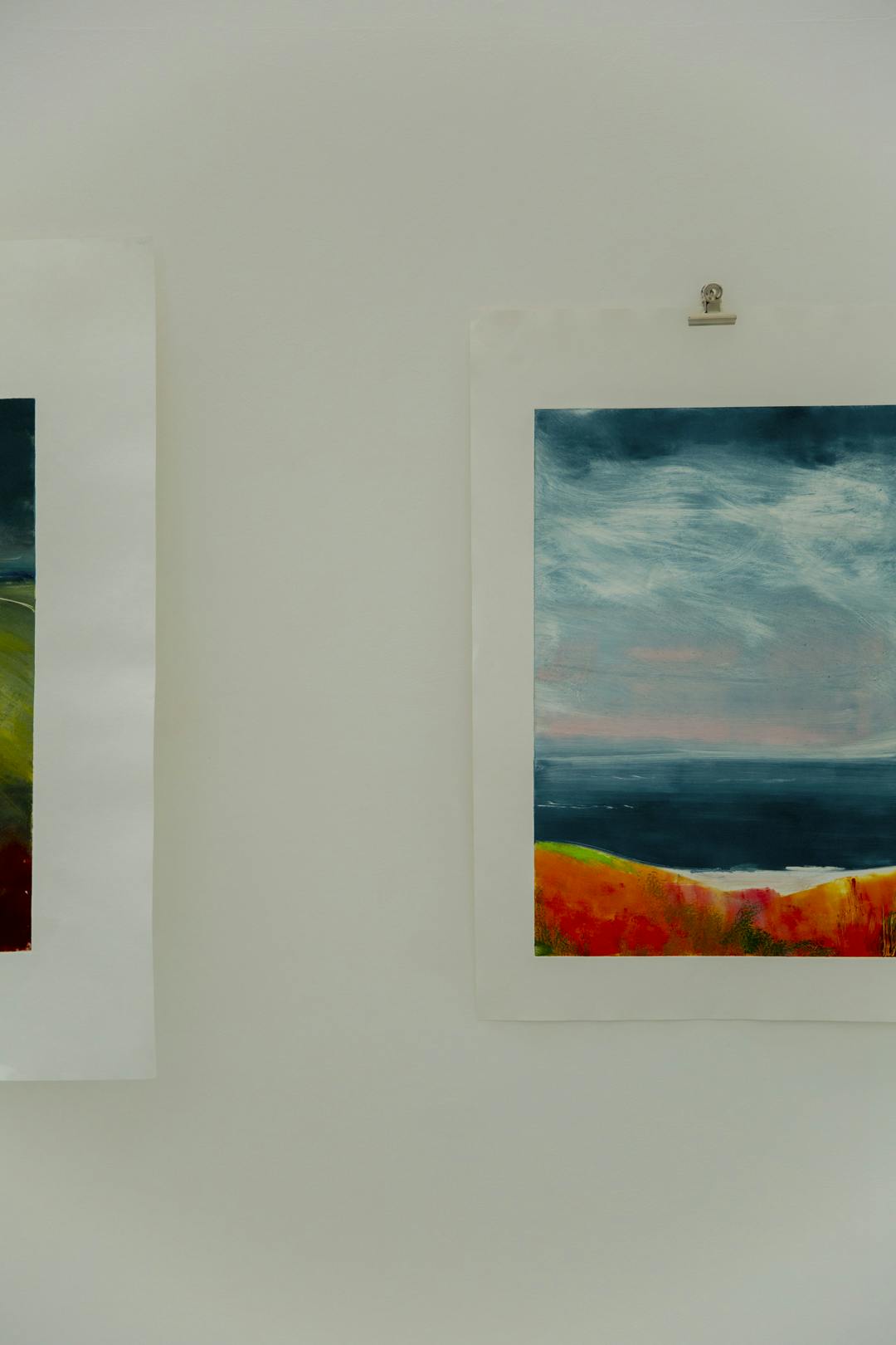 Image shows Martine's work hung up in the gallery space with one half of a painting on the left and another half of a painting on the right. The painting features blues and orange.