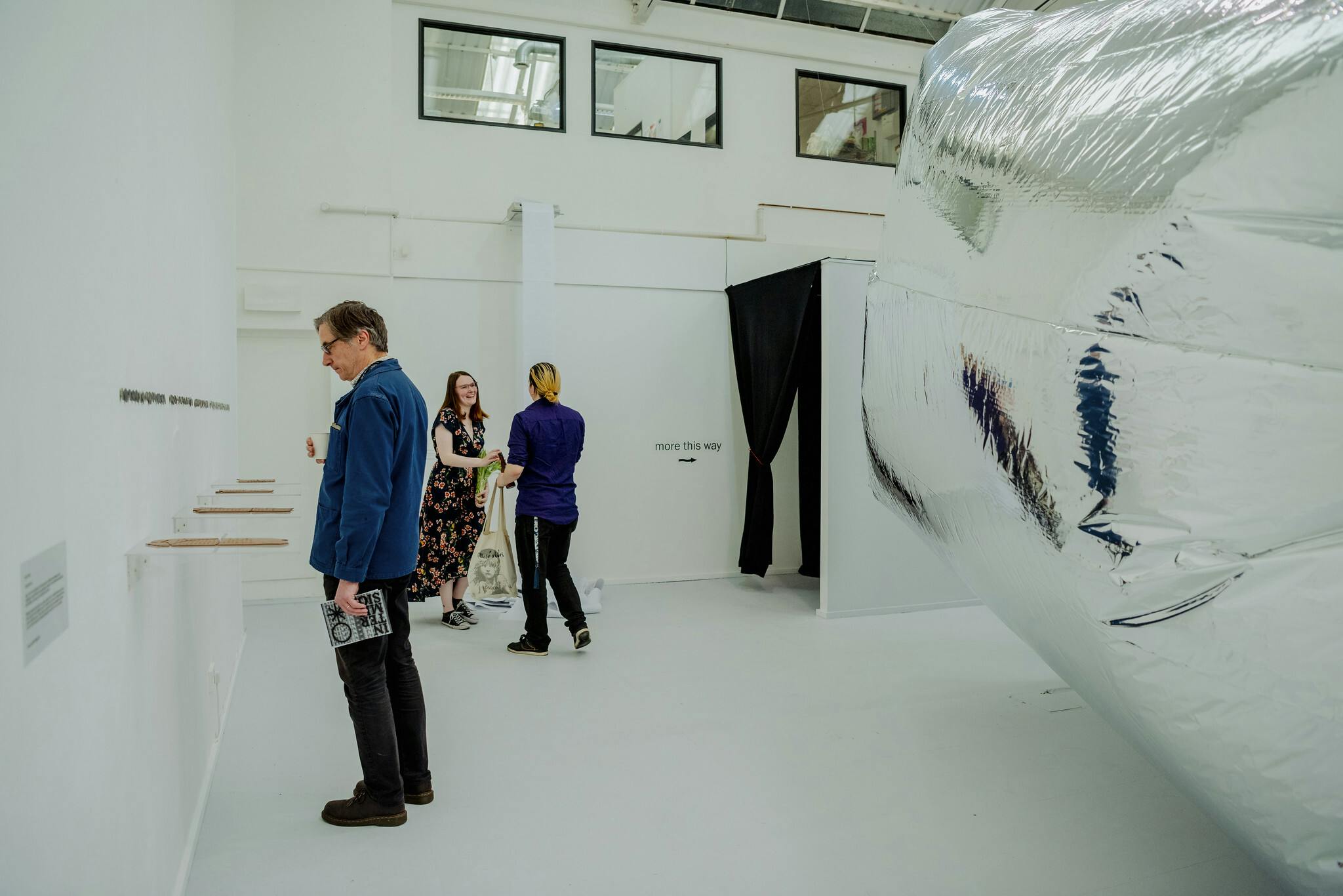 Three people stand in an exhibition space looking at work including clay pieces in glass boxes and a large inflatable silver work that fills the right of the photo