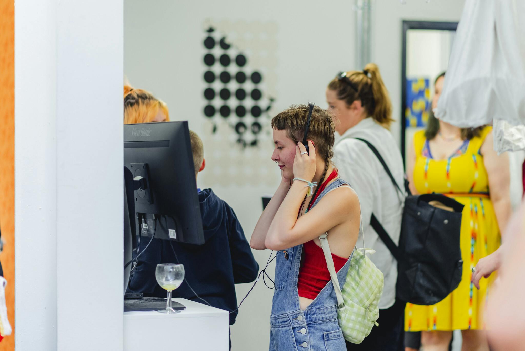 A young woman in dungarees wears headphones whilst watching an animation on a screen within a busy exhibition space