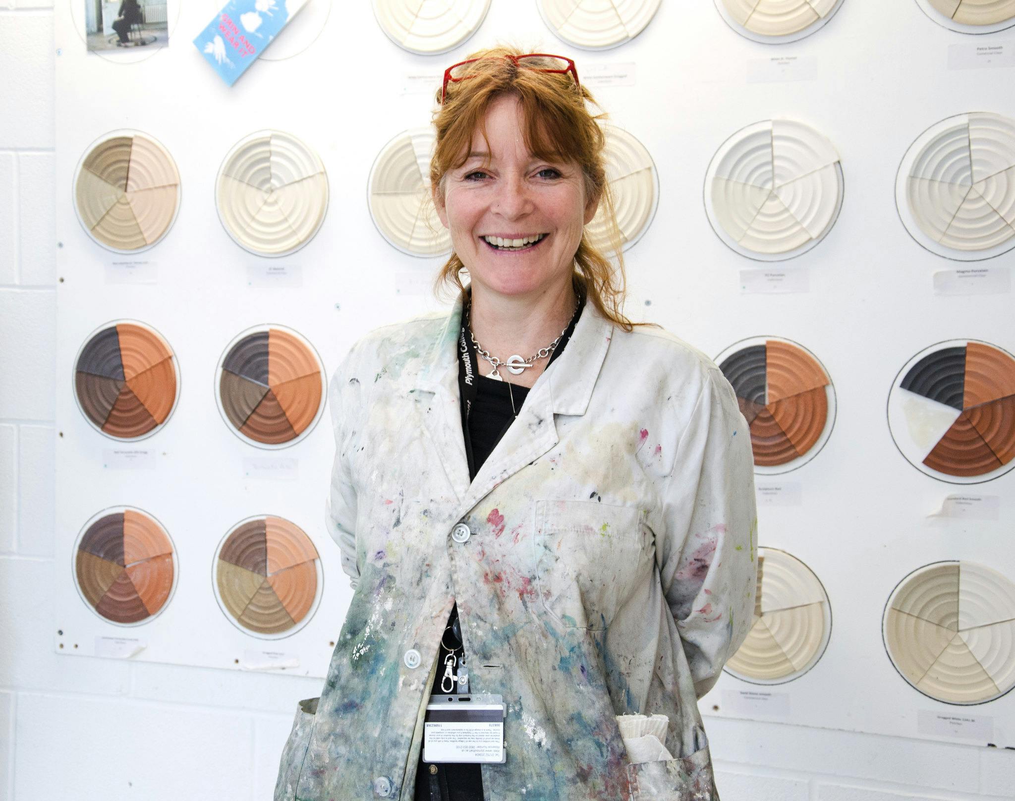 Crafts student Jo Tyler smiles at the camera in our ceramics studios with a wall of test tiles behind her