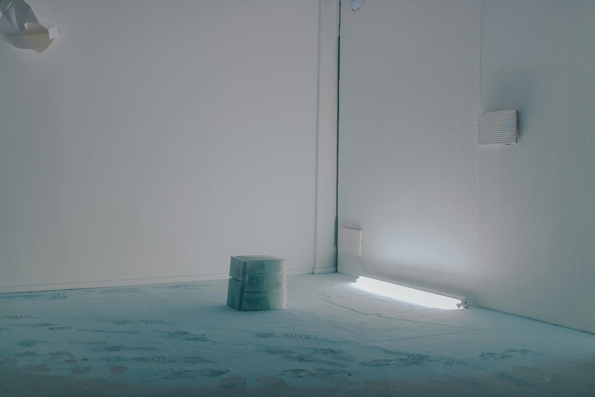 An exhibited piece featuring a white cube in the corner of a room lit with a white strip light and surrounded by white walls