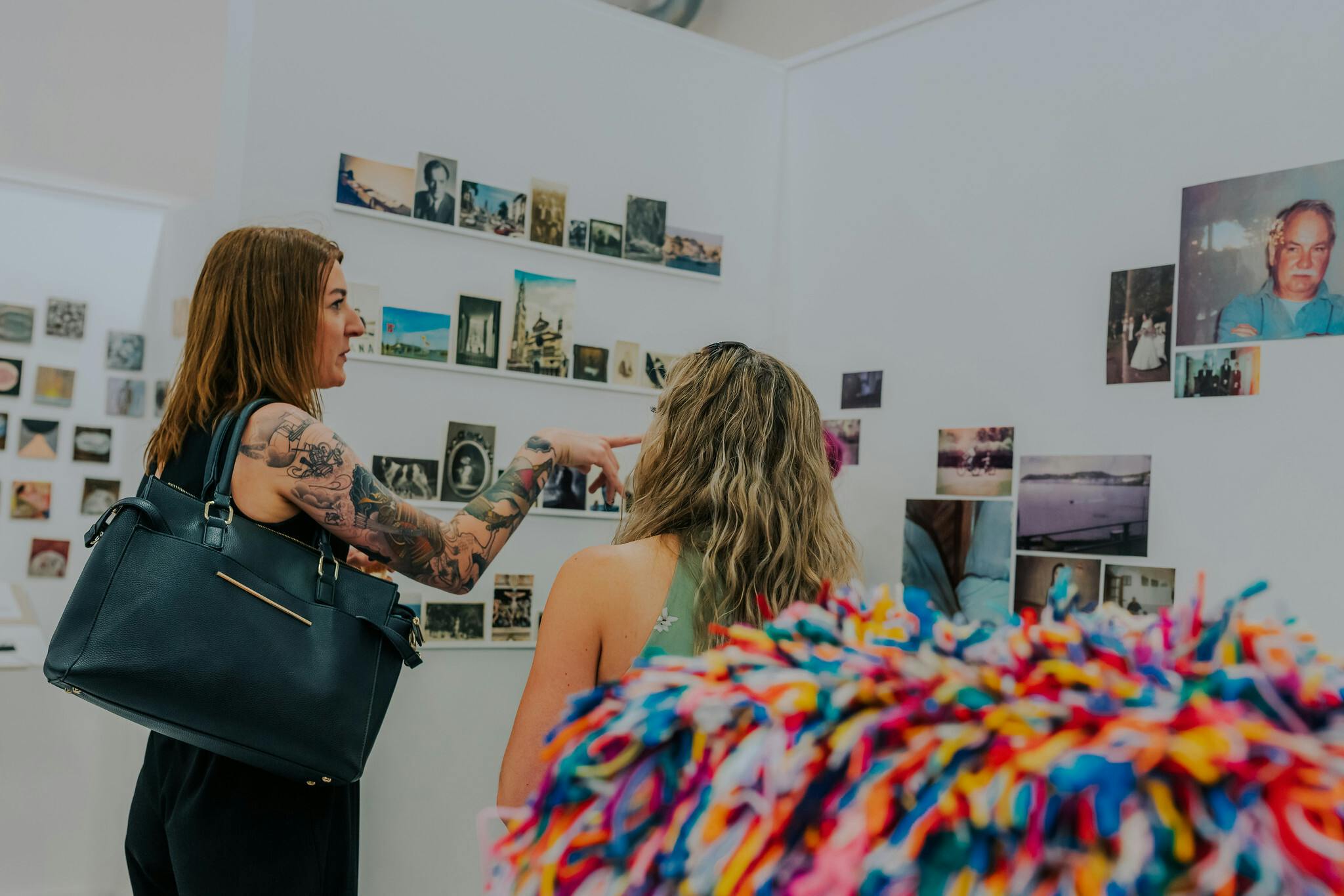 A heavily tattooed woman looking and pointing at photographs on the wall while a blonde woman with wavy hair looks on. In the foreground there is a colourful rainbow textile ball.