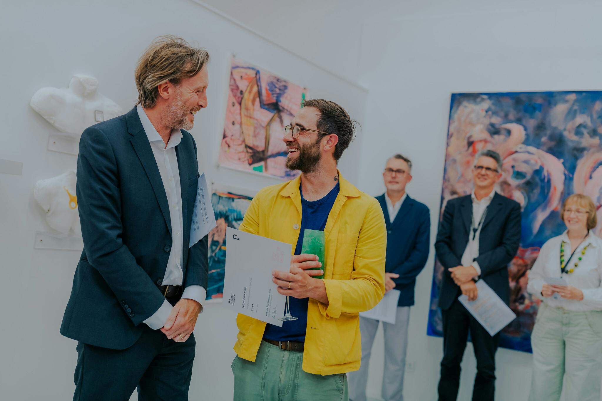 Vice Chancellor Paul Fieldsend-Danks smiling at MA student William Luz while Dr Stephen Paige, Prof Stephen Felmingham and Board of Governors Chair Sue Brownlow watch on in the background surrounded by colourful paintings