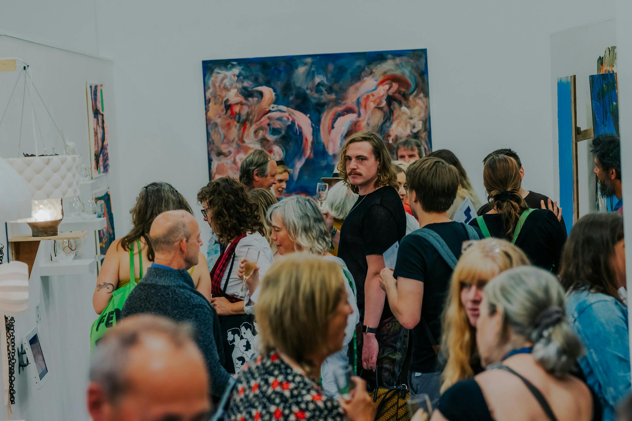 A crowd of people in an exhibition space with a large swirling painted canvas in the background and other exhibited items to the left and right