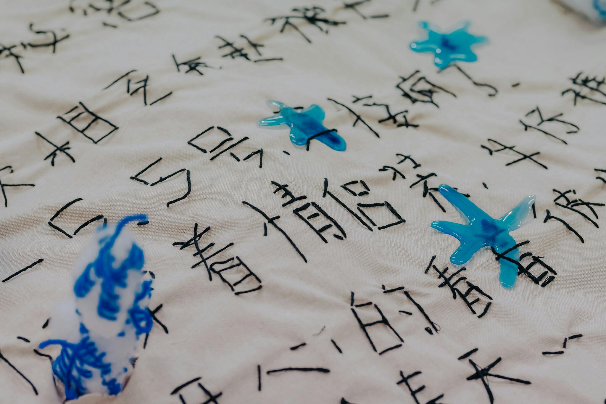 Embroidered lettering on a pale fabric with drops of bright blue wax throughout the image