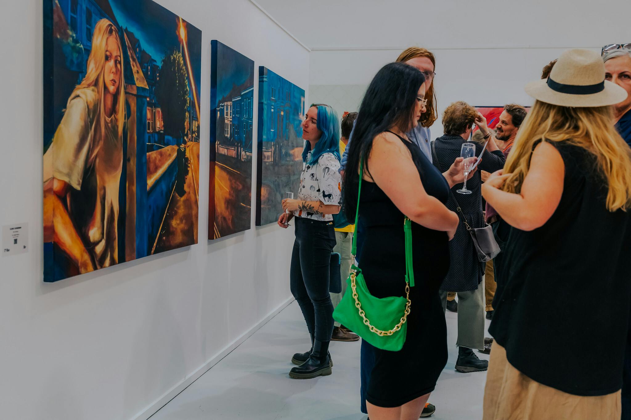 People in the foreground at an exhibition and in the background a woman with blue hair looks on at a trio of blue and orange paintings of a woman