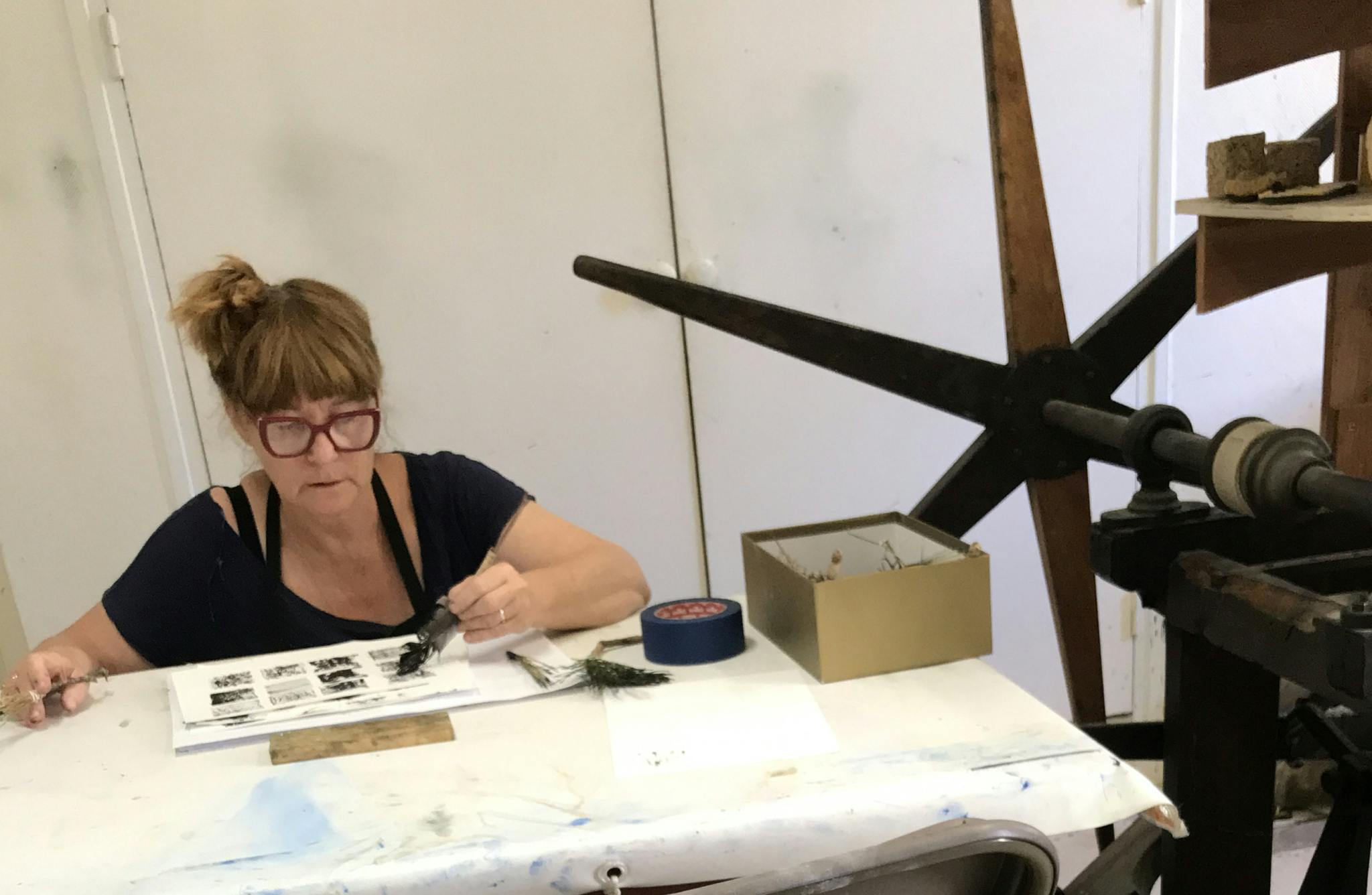 Jo Tyler brushes black ink on paper on a table next to a large printing press