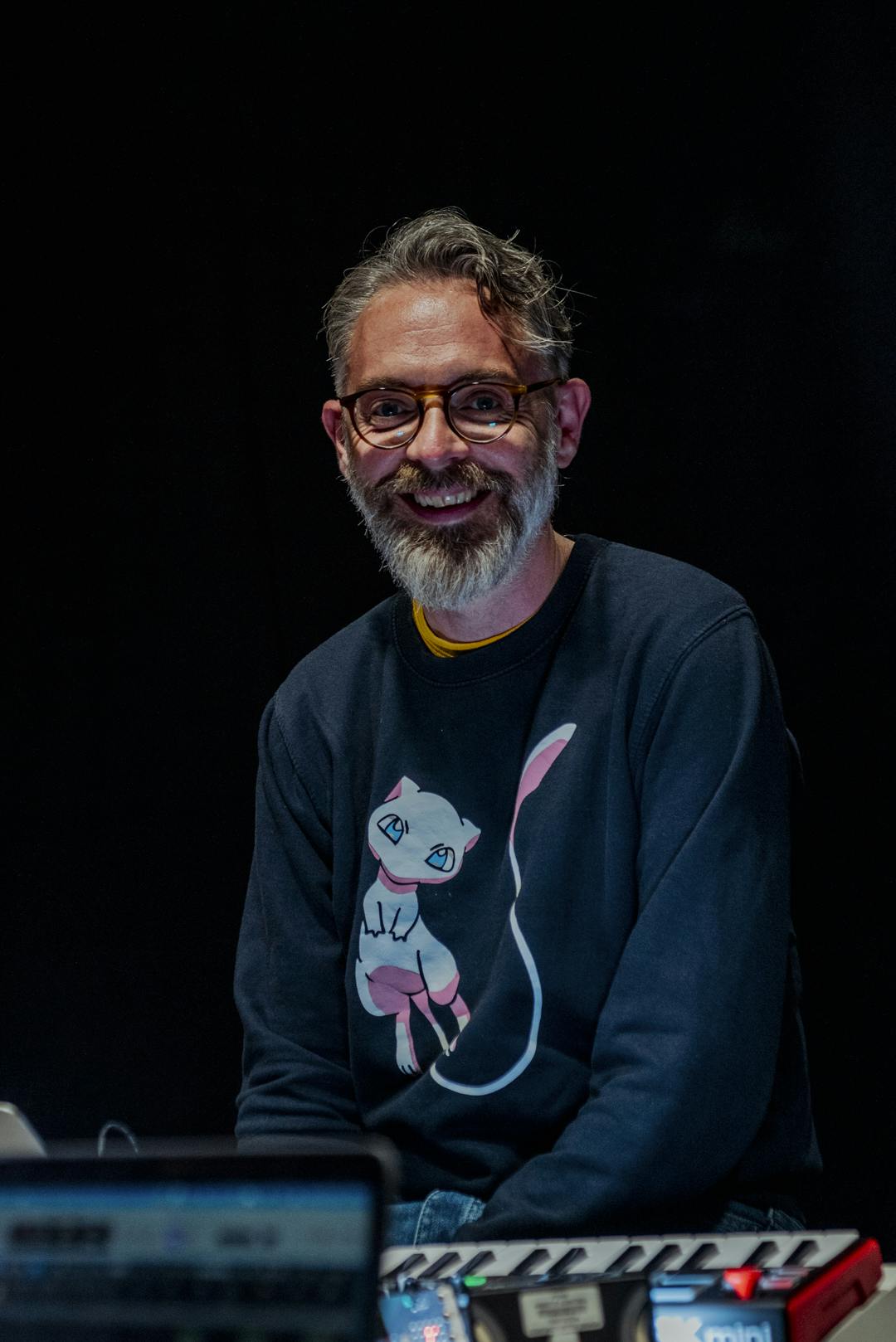 Neil Rose, Sound Arts Lecturer, wears a long sleeve black sweatshirt and glasses as he smiles whilst working on the sound mixing desks on a table in front of him